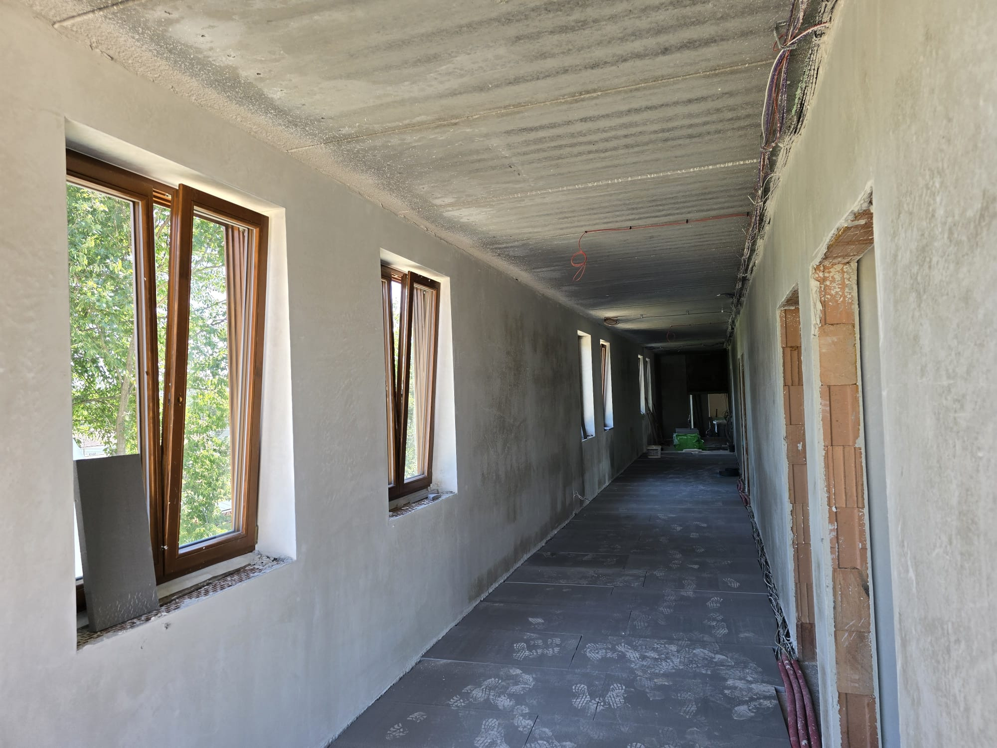 Korytarz w stanie surowym po nałożeniu tynków maszynowych. Widoczne okna z drewnianą stolarką, ślady na podłodze i instalacje pod sufitem.