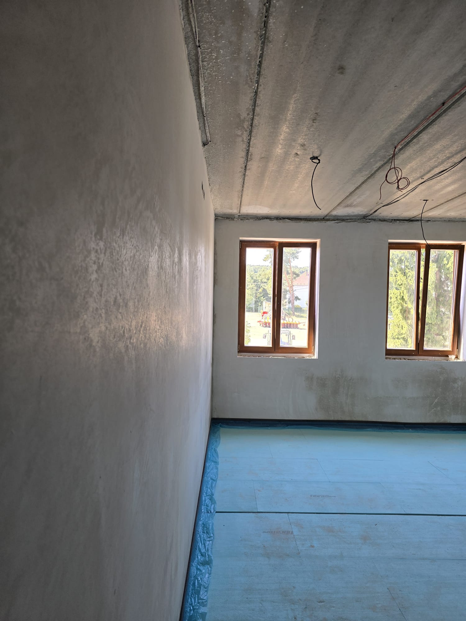 Gładka, jasnoszara ściana po tynkowaniu maszynowym, z widocznymi dwoma oknami i zabezpieczoną podłogą. Surowy betonowy sufit z instalacją elektryczną.