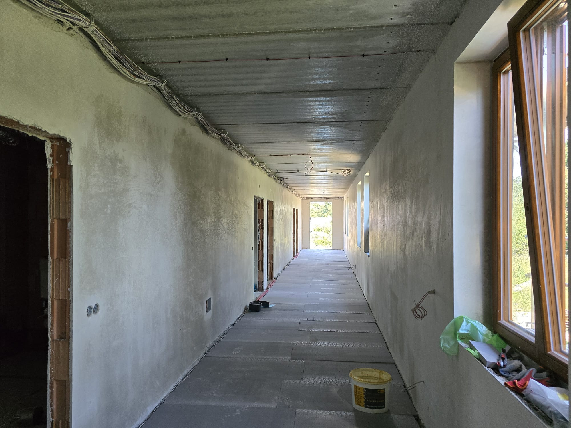 Korytarz w trakcie prac wykończeniowych: ściany po tynkowaniu, widoczne instalacje elektryczne pod sufitem, podłoga przygotowana pod wylewkę, okno z widokiem na zieleń.