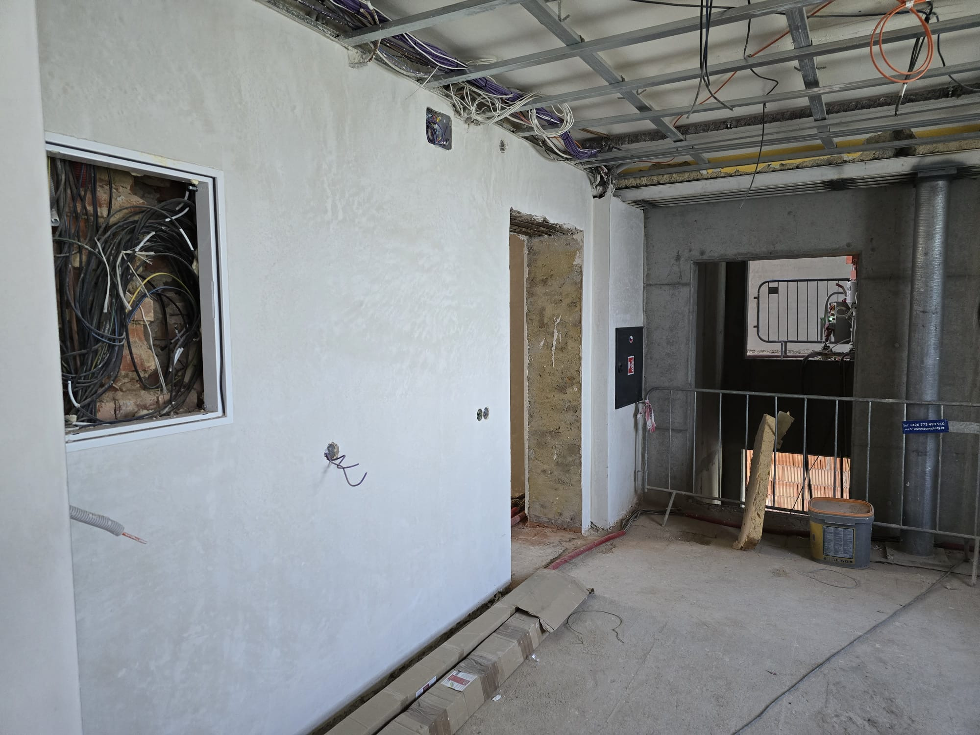 Wnętrze w trakcie prac wykończeniowych: ściana z nałożonym tynkiem, widoczne instalacje elektryczne w ścianie i pod sufitem, surowy stan podłogi.