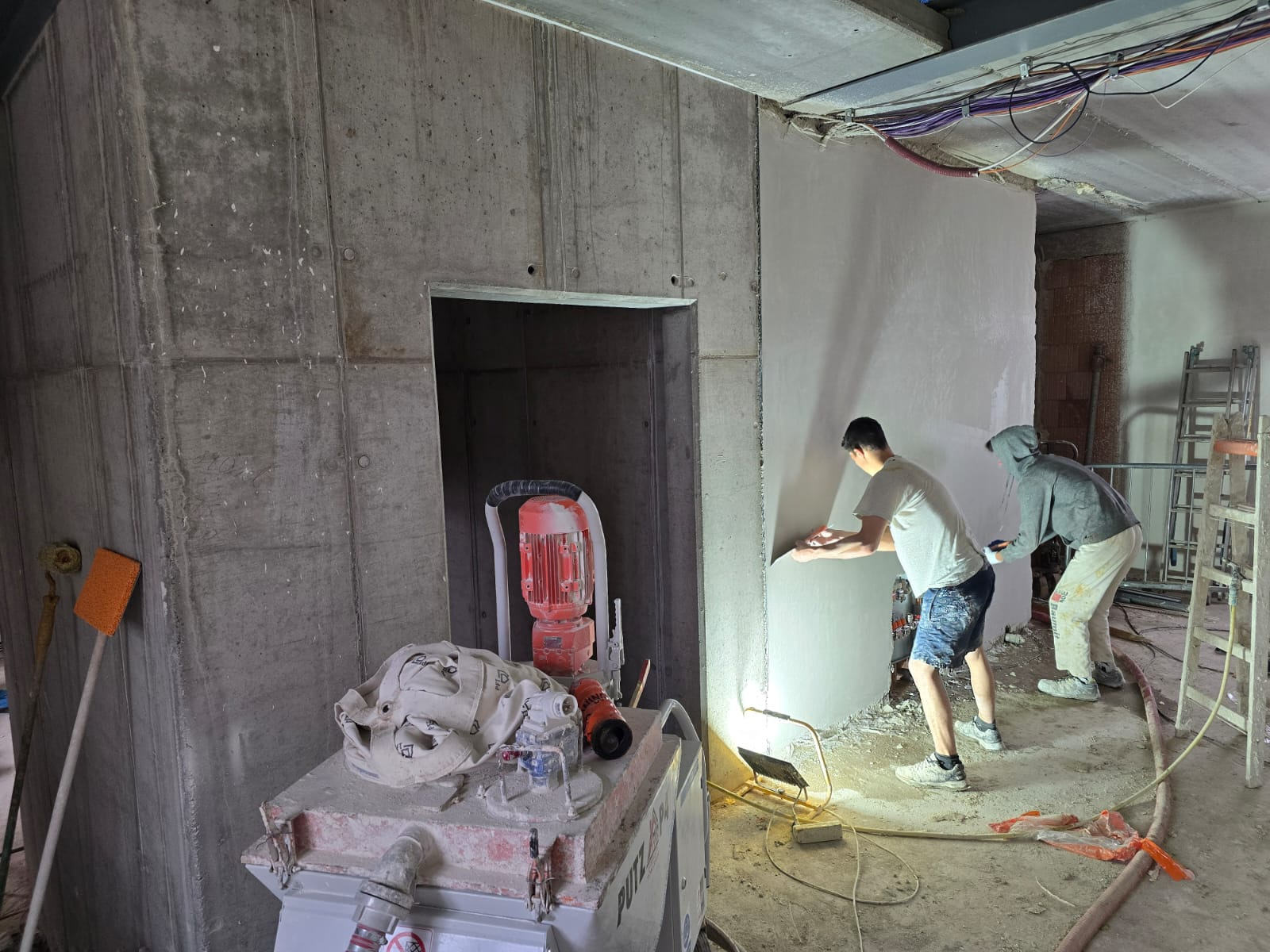 Praca tynkarska w surowym wnętrzu: dwóch mężczyzn aplikuje tynk na ścianę. Widoczna maszyna do tynkowania, oświetlenie robocze i betonowe ściany w tle.