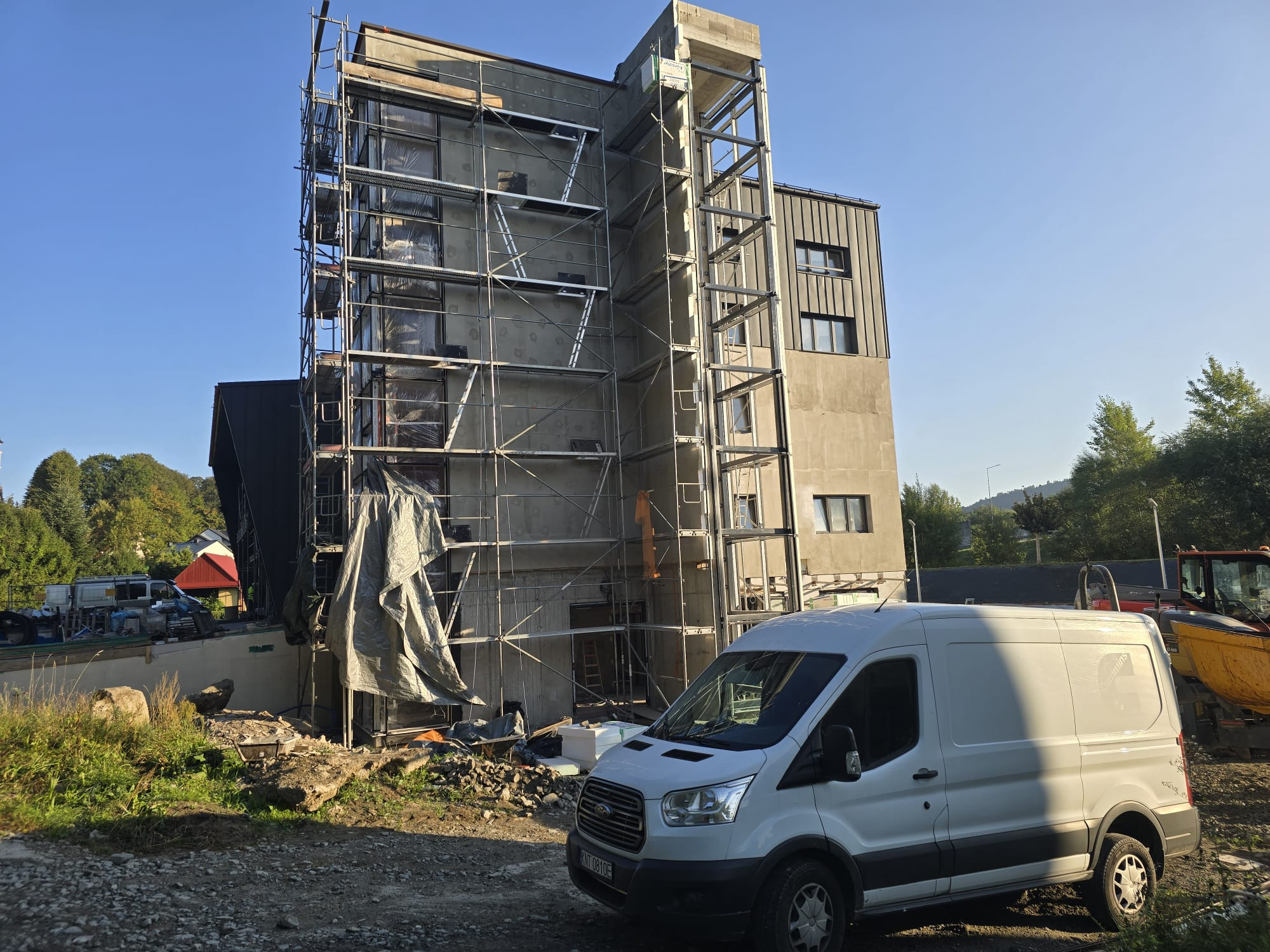 Budynek w trakcie tynkowania, z rusztowaniem i osłoniętą plandeką częścią elewacji. Biały bus firmowy na pierwszym planie. Widok na plac budowy w słoneczny dzień.
