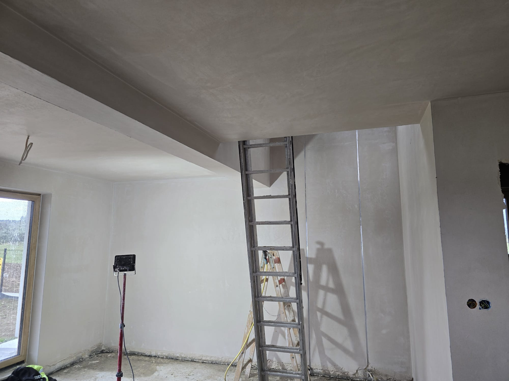 Wnętrze po tynkowaniu maszynowym: gładkie, jasne ściany, drabina oparta o ścianę, widoczne okno i lampa budowlana. Świeżo położone tynki.