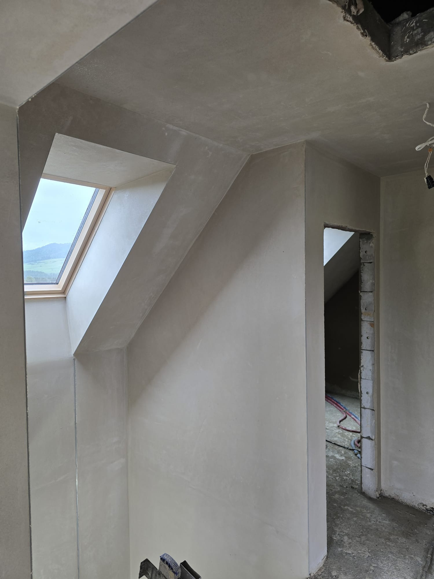 Jasne wnętrze po nałożeniu gładzi, widoczne okno dachowe i przejście do innego pomieszczenia. Ściany i sufit pokryte świeżym tynkiem.