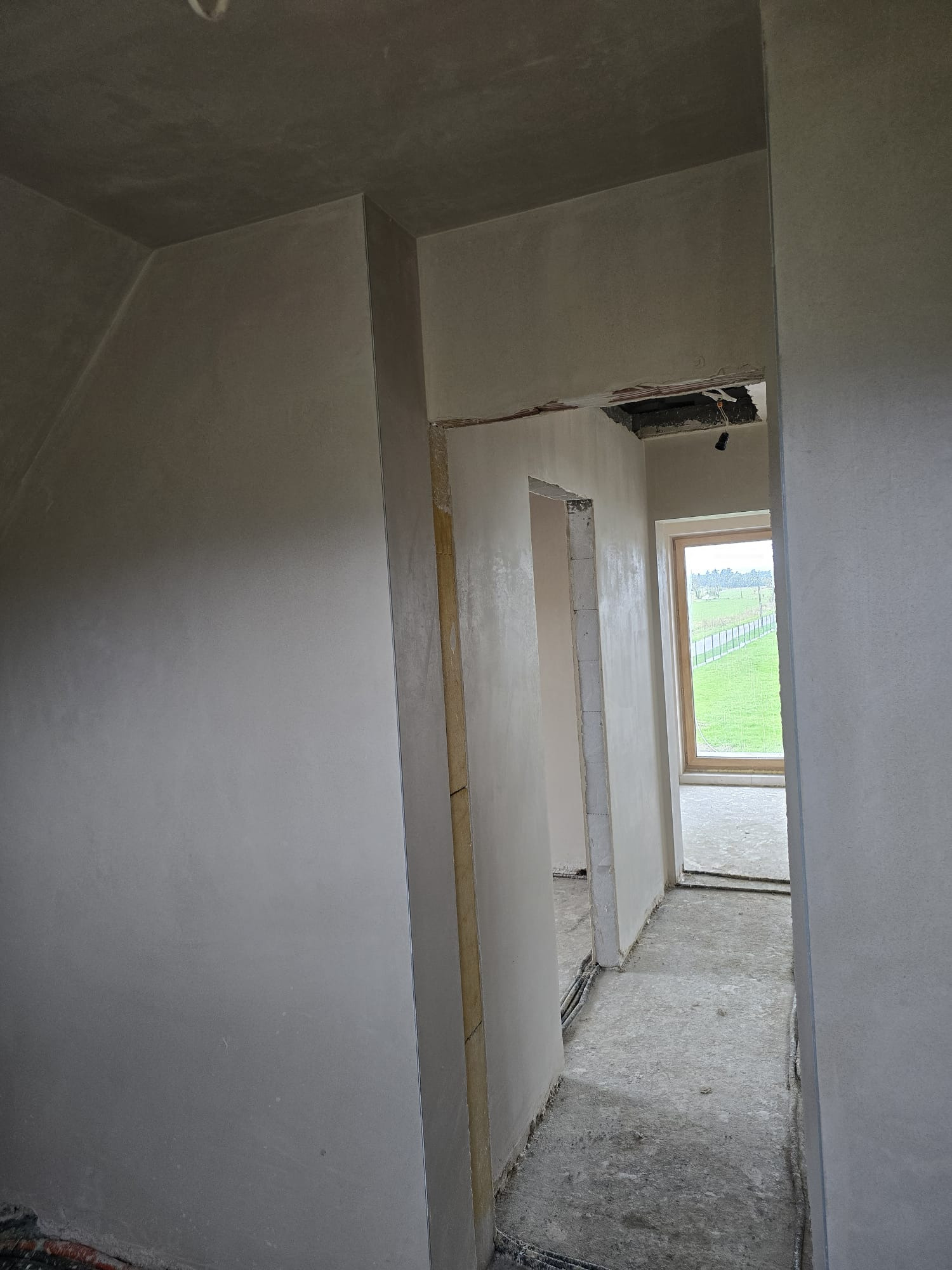 Wnętrze domu po tynkowaniu maszynowym: gładkie ściany, widoczne kable w suficie, okno z widokiem na zewnątrz. Szare odcienie, surowy stan wykończenia.