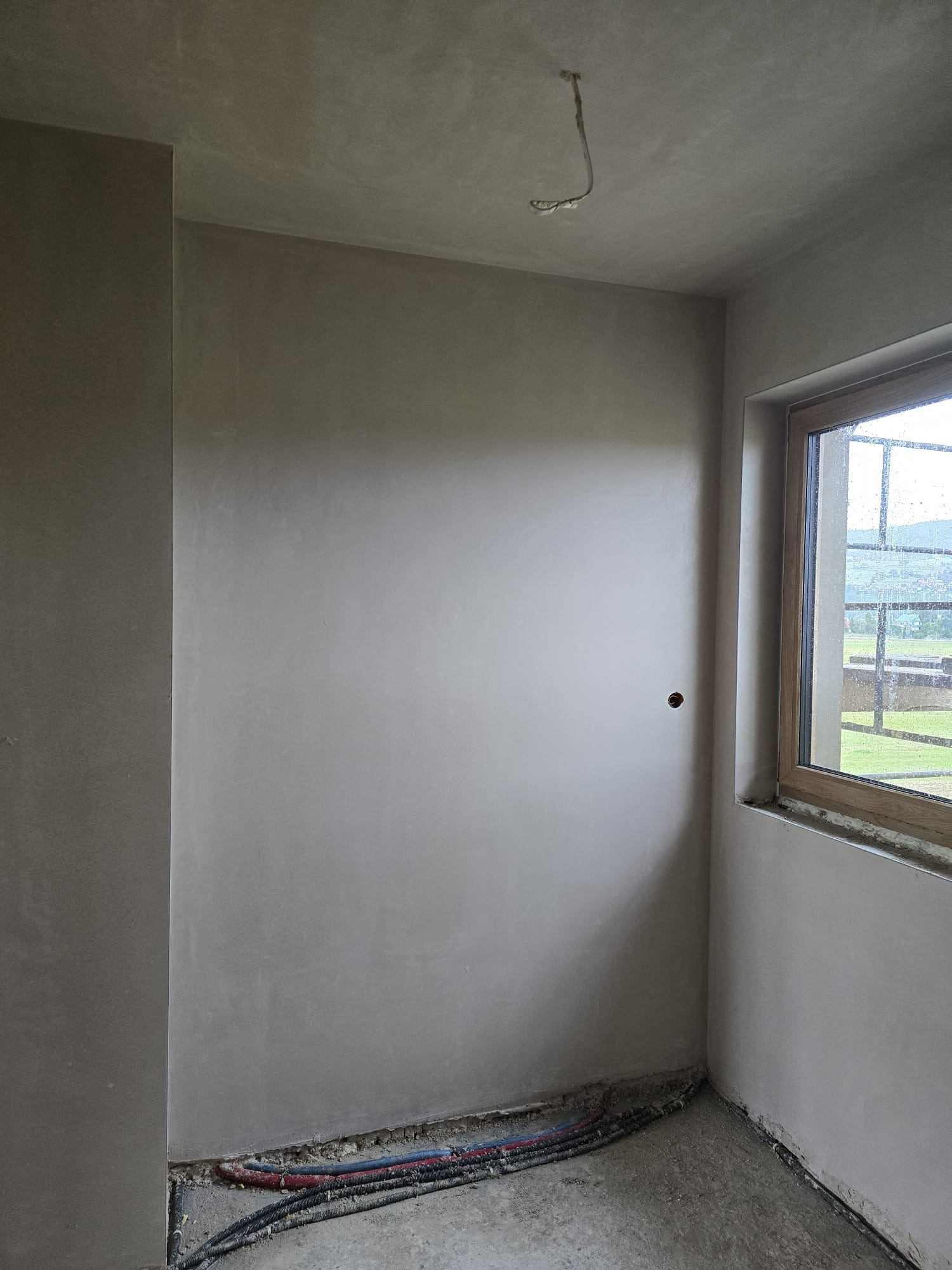 Gładkie, szare ściany po tynkowaniu maszynowym w nowym wnętrzu. Widoczne kable przy podłodze i okno z widokiem na zieleń. Punkt elektryczny na ścianie.