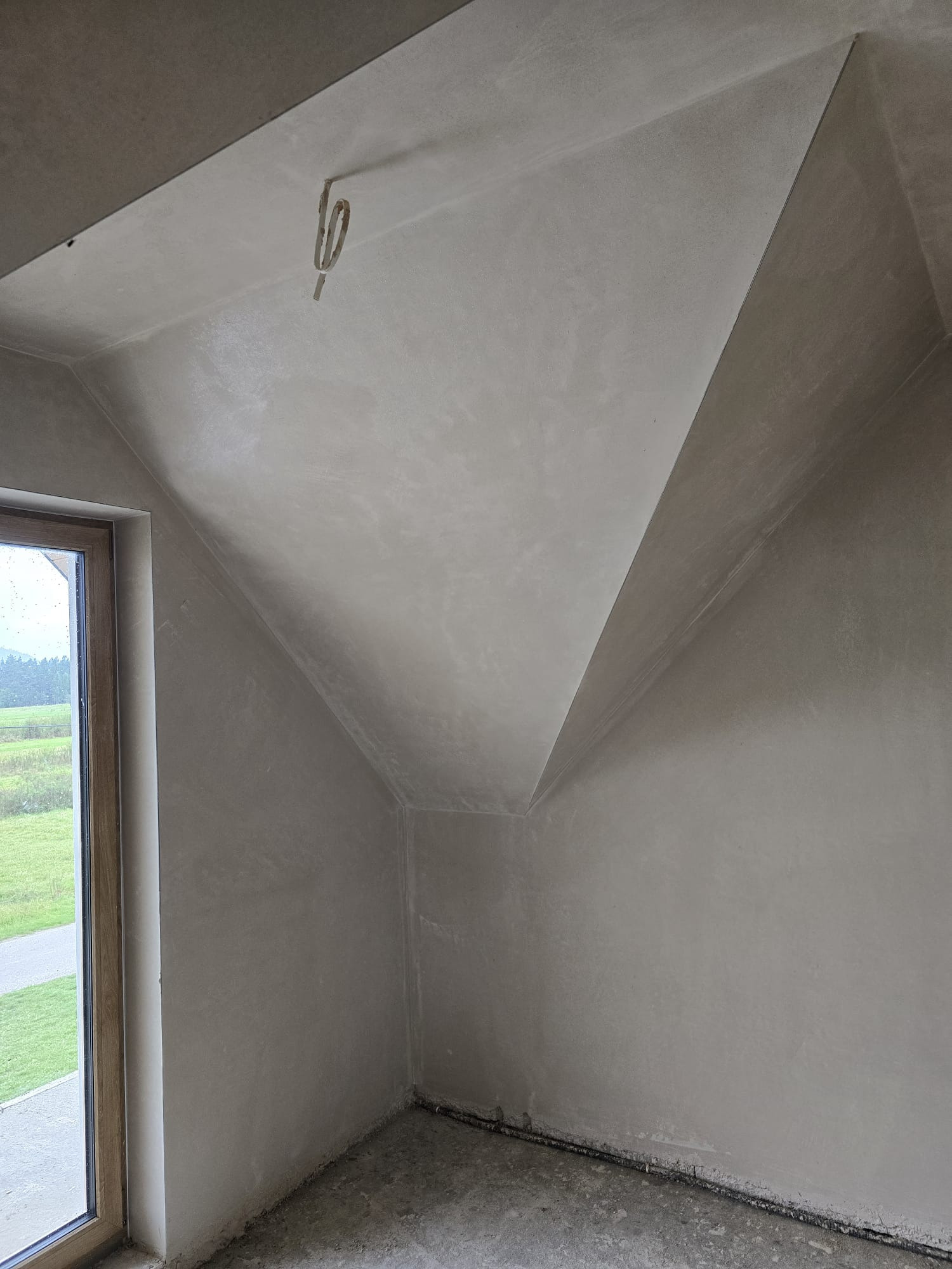 Szare, gładkie ściany poddasza po tynkowaniu maszynowym, widoczne okno z widokiem na zewnątrz i wystający kabel elektryczny z sufitu.