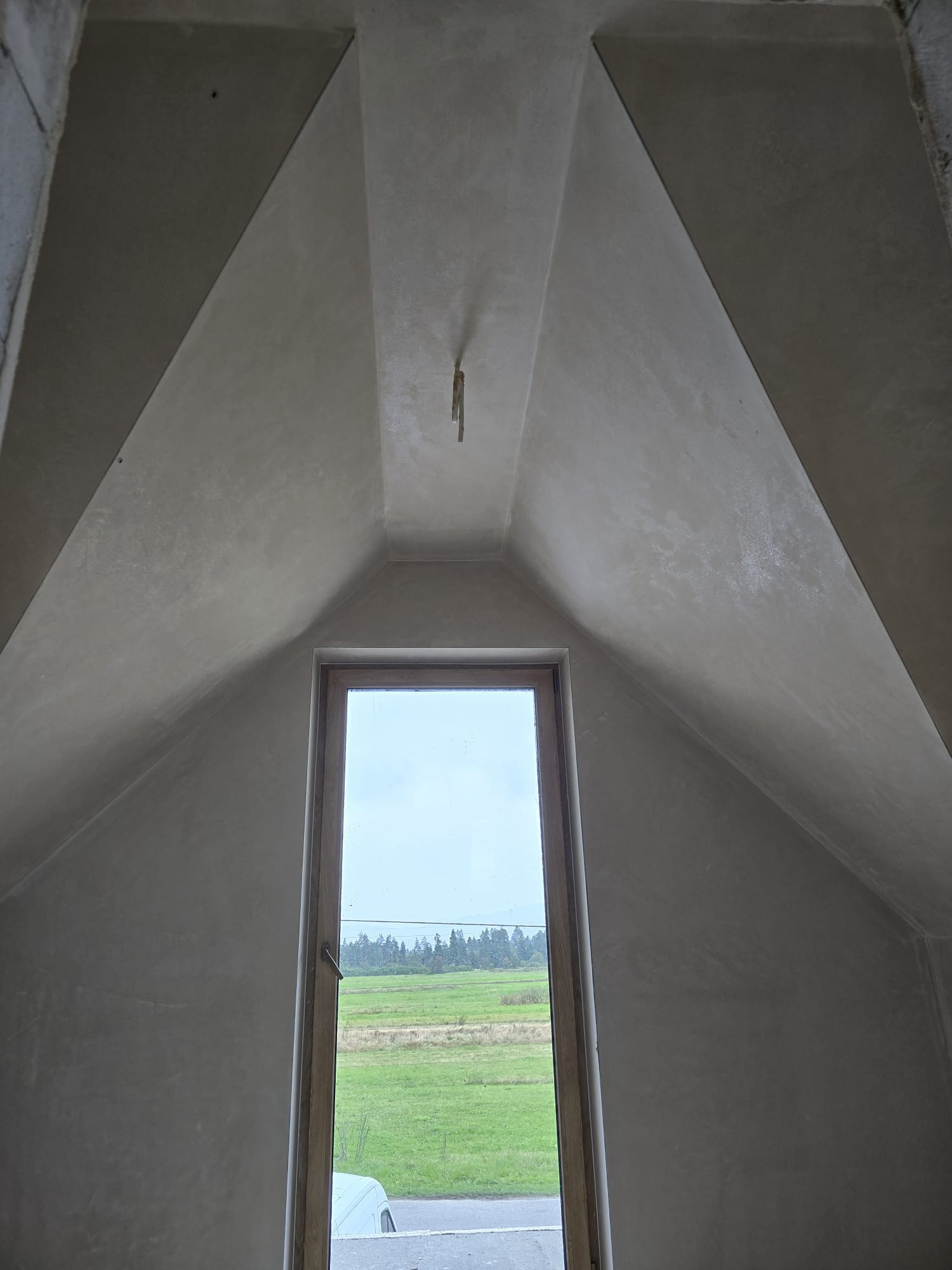 Poddasze z oknem po nałożeniu tynków. Widoczne gładkie, szare ściany i sufit ze skosami. Okno z widokiem na zielone pole i las w oddali.