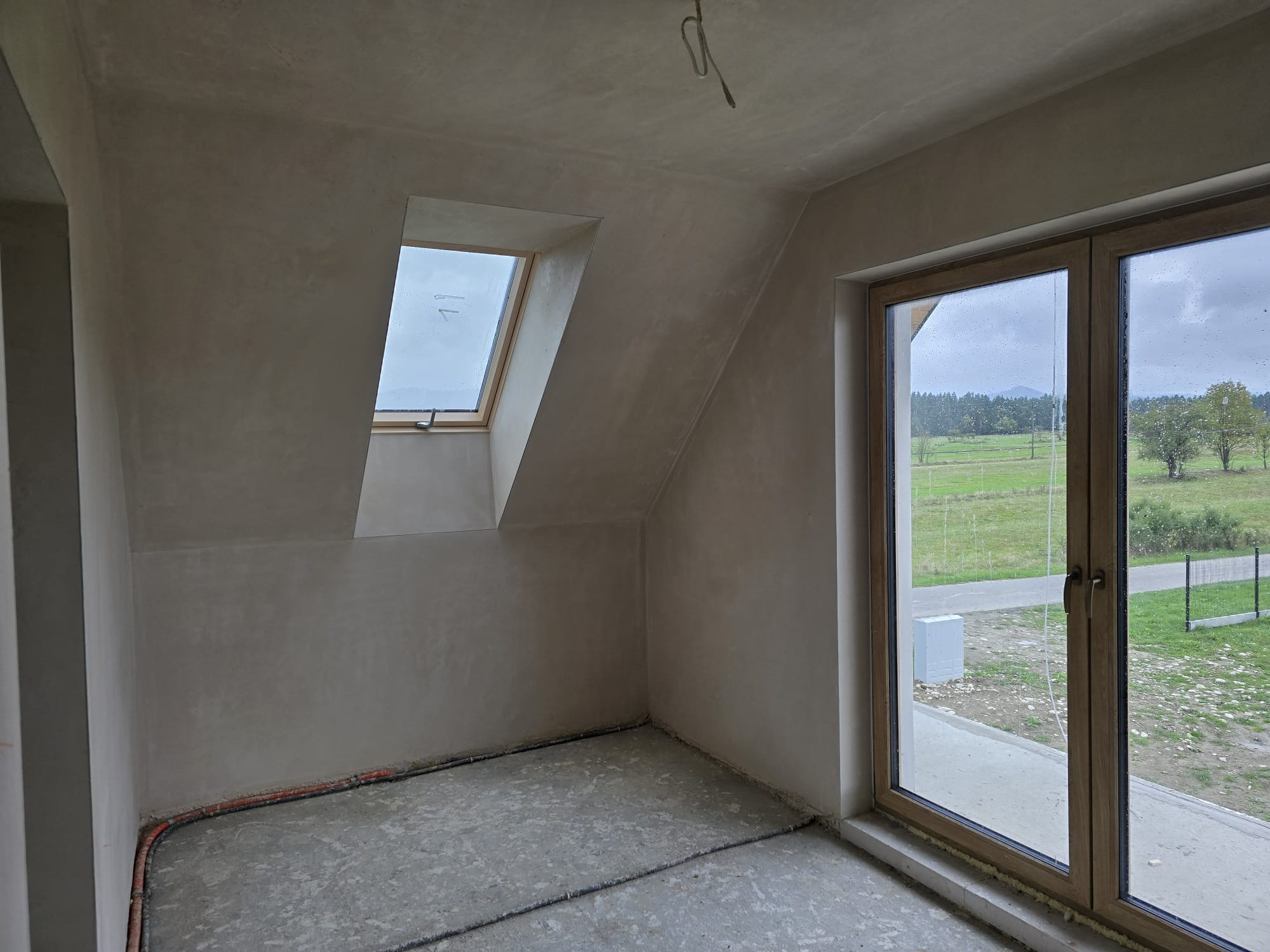 Pokój na poddaszu po nałożeniu tynków maszynowych. Widoczne okno dachowe i duże okno balkonowe z widokiem na zewnątrz. Ściany i sufit w jasnym kolorze, świeżo po tynkowaniu.