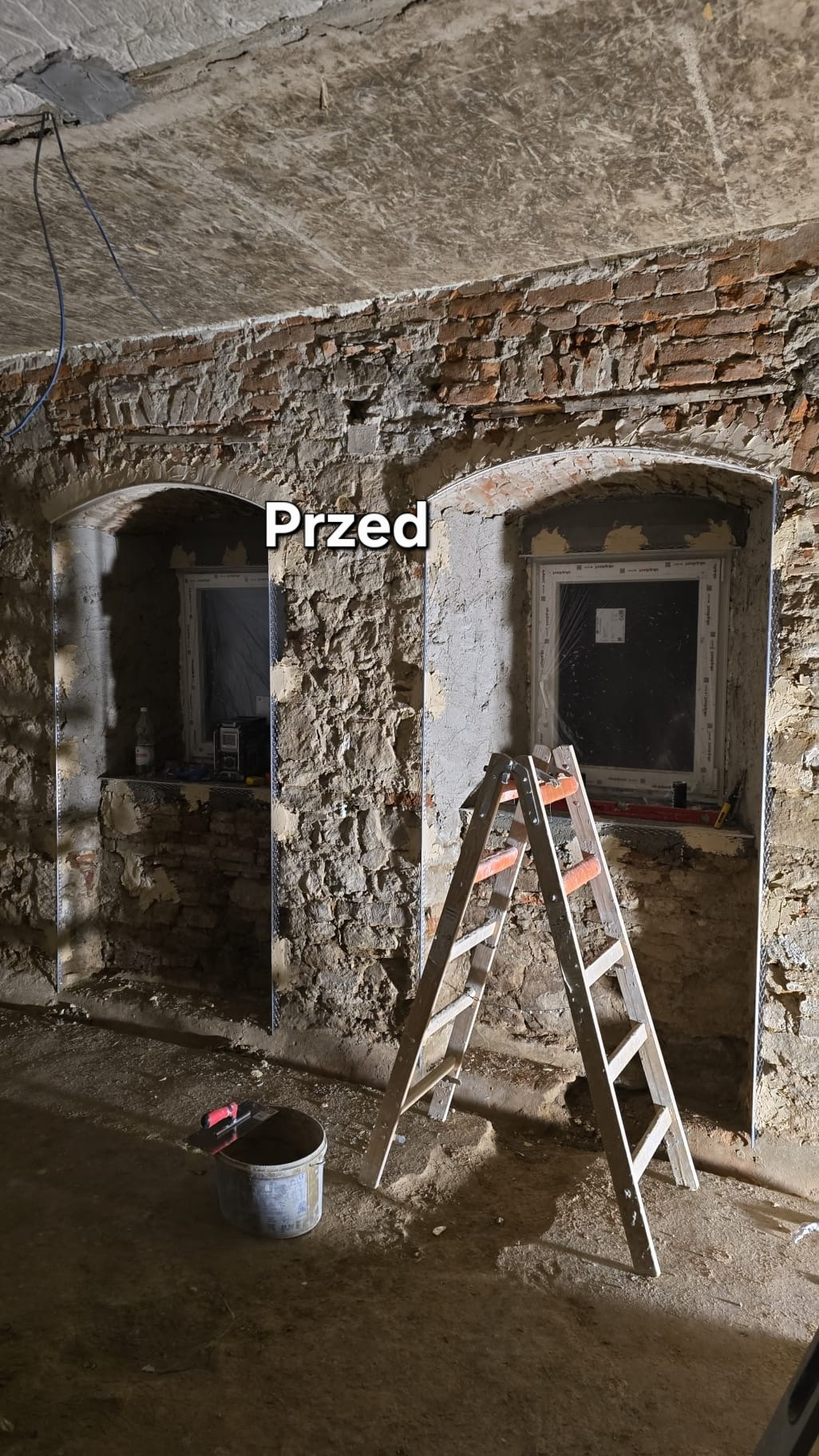 Remont piwnicy: dwa okna przygotowane do tynkowania, widoczna stara cegła, siatka tynkarska, drabina i kubeł. Etap 'przed' tynkowaniem maszynowym.
