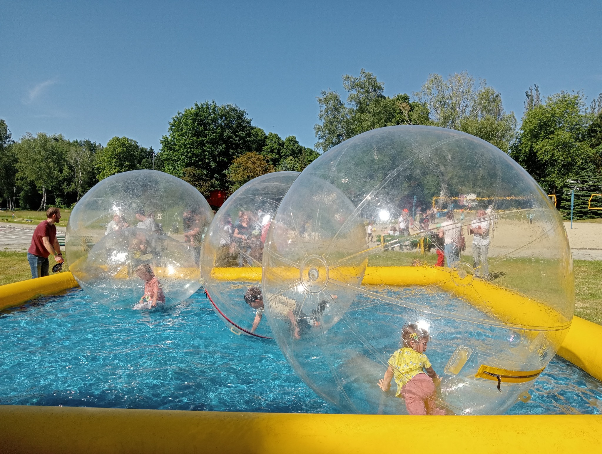 Atrakcja wodna: dzieci wewnątrz przezroczystych kul unoszących się na basenie otoczonym żółtymi dmuchanymi elementami, w tle drzewa i niebieskie niebo.