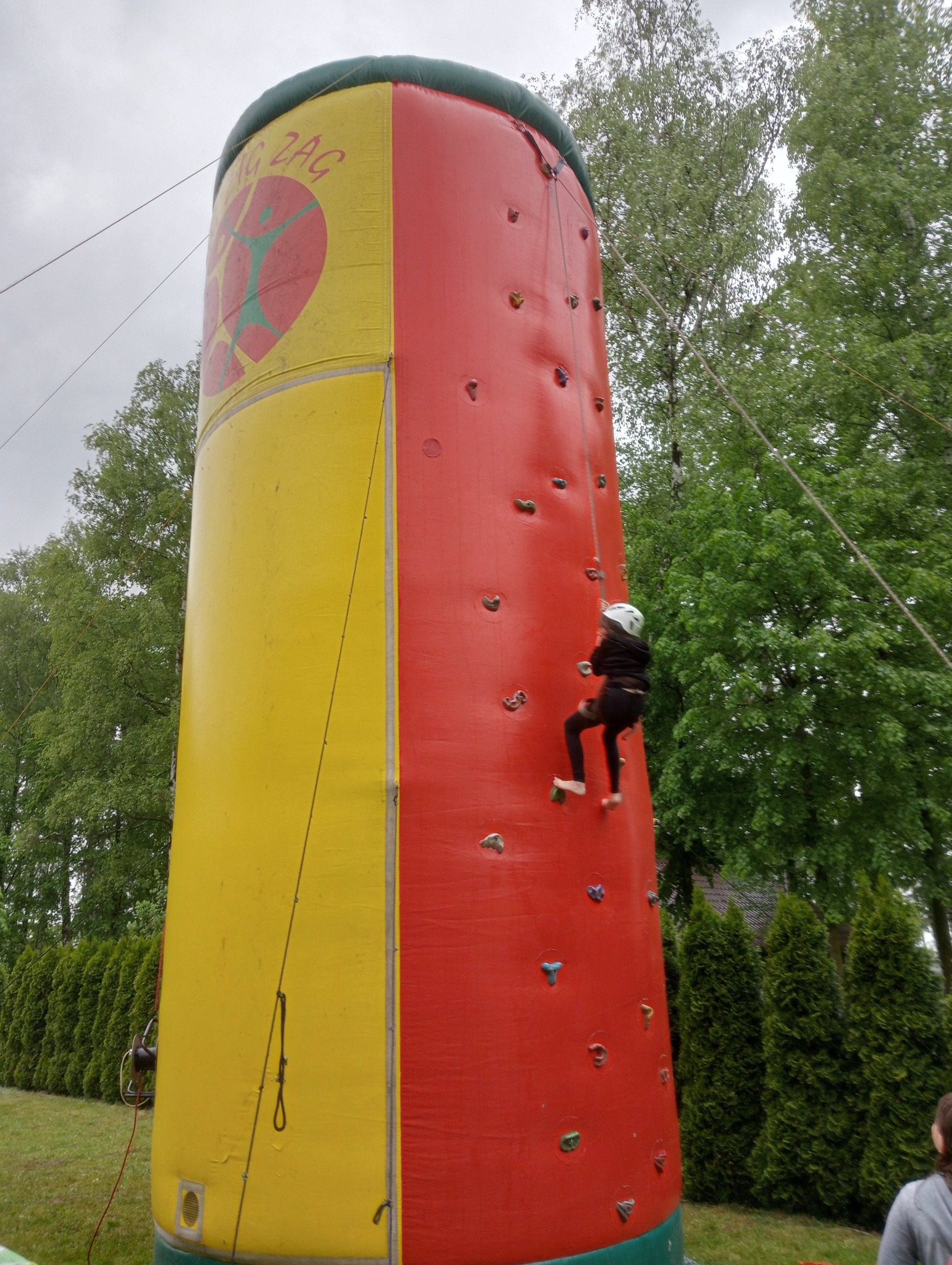 Dziecko w kasku wspina się na dmuchaną ściankę wspinaczkową, żółto-czerwoną, z logo Zig Zag. W tle zieleń drzew i krzewów, pochmurne niebo.