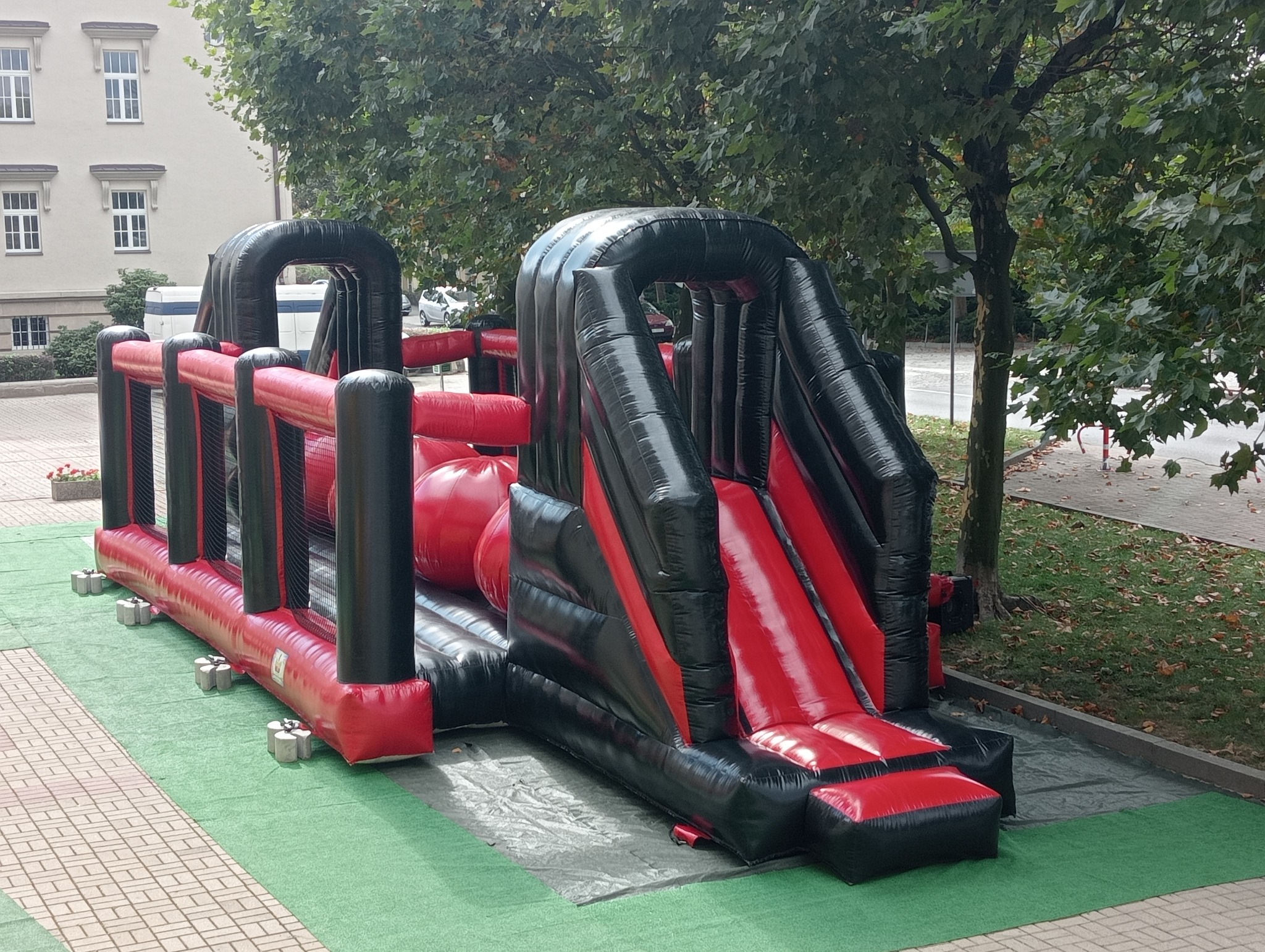 Czarno-czerwony dmuchany tor przeszkód z tunelem i zjeżdżalnią na zielonej trawie, z budynkiem i drzewami w tle. Widoczne elementy mocujące.