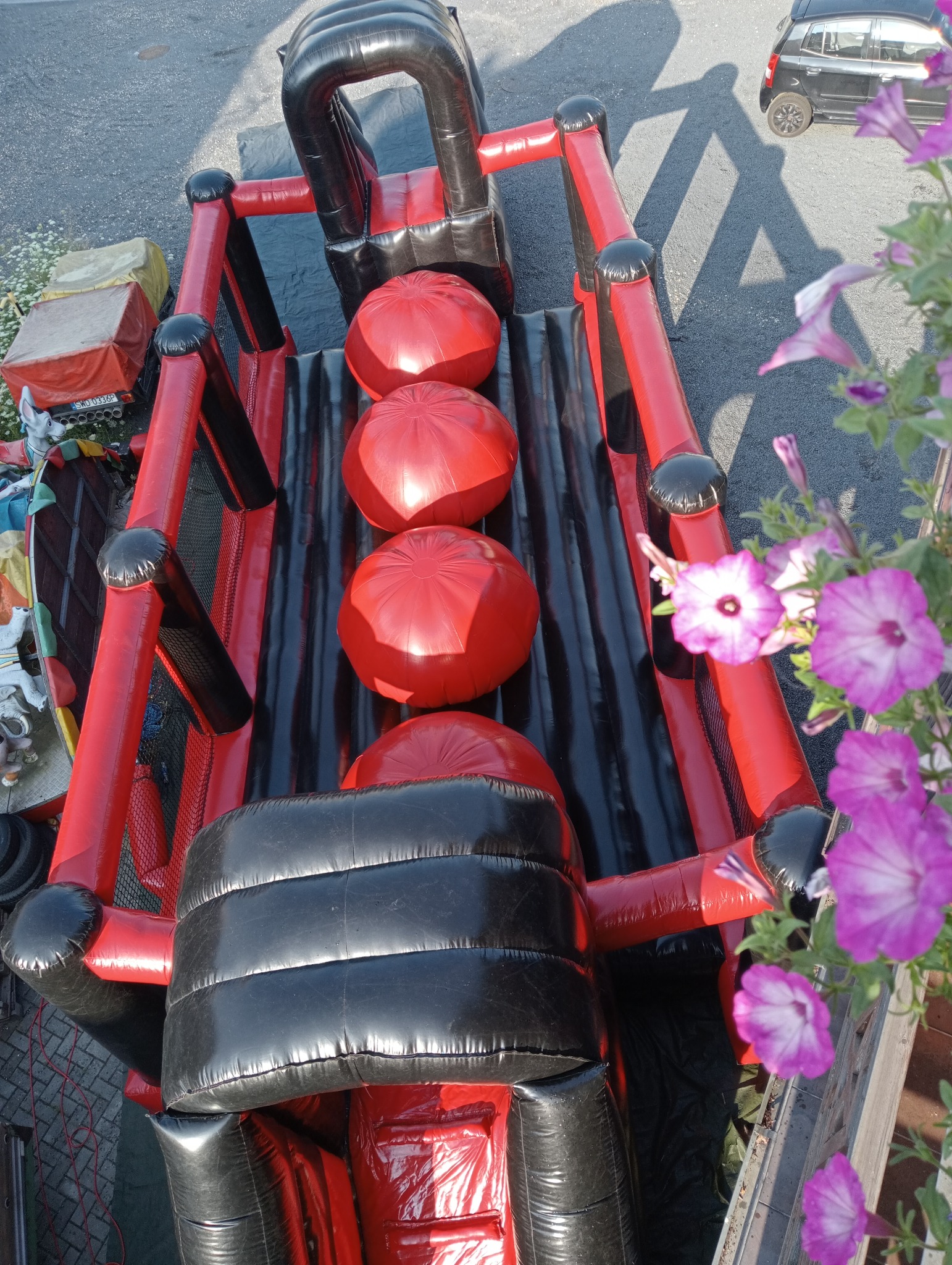 Widok z góry na czerwono-czarny dmuchany tor przeszkód z trzema czerwonymi kulami, częściowo zasłonięty przez fioletowe kwiaty i z widocznym fragmentem samochodu.