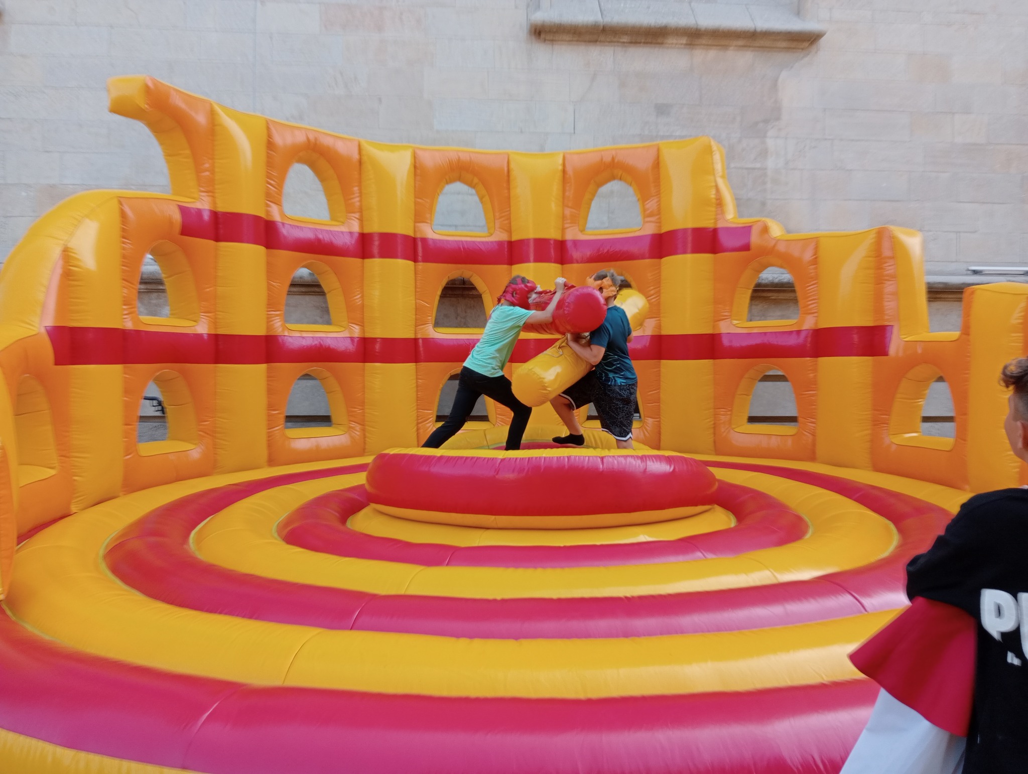 Dwie osoby w strojach ochronnych walczą na dmuchanym ringu w kolorach żółto-czerwonych. W tle dmuchana konstrukcja z otworami, przypominająca arenę.