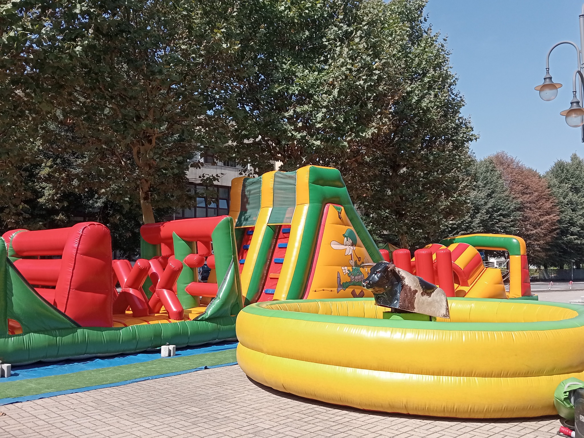 Kolorowy park dmuchany z bykiem rodeo na placu, otoczony zielenią drzew i budynkami w tle. Dmuchana zjeżdżalnia z grafiką i tor przeszkód.