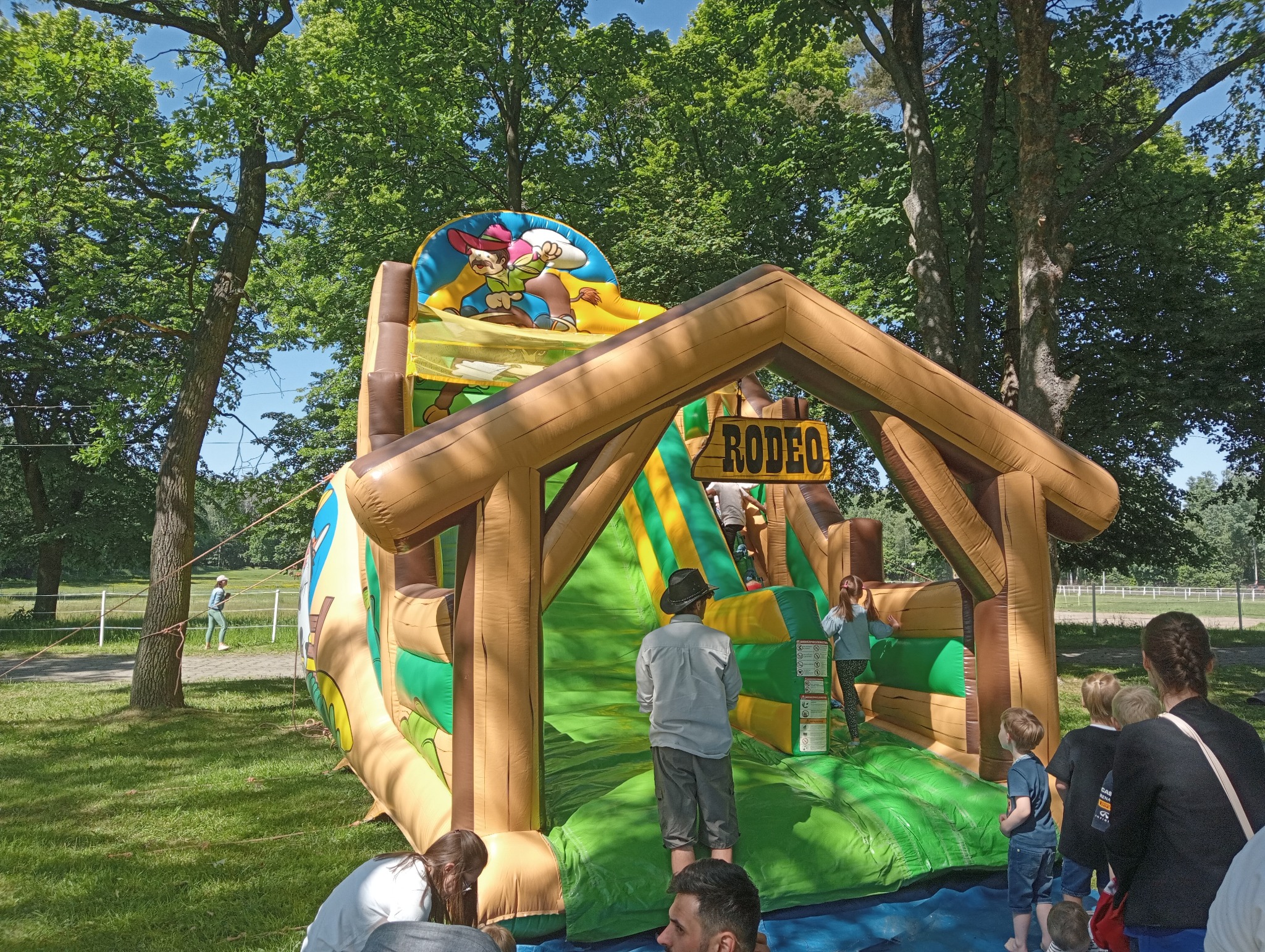 Dmuchany zamek w stylu rodeo z dziećmi w parku, otoczony drzewami i zielenią. Atrakcja plenerowa z motywem kowbojskim, idealna na imprezy dla dzieci.
