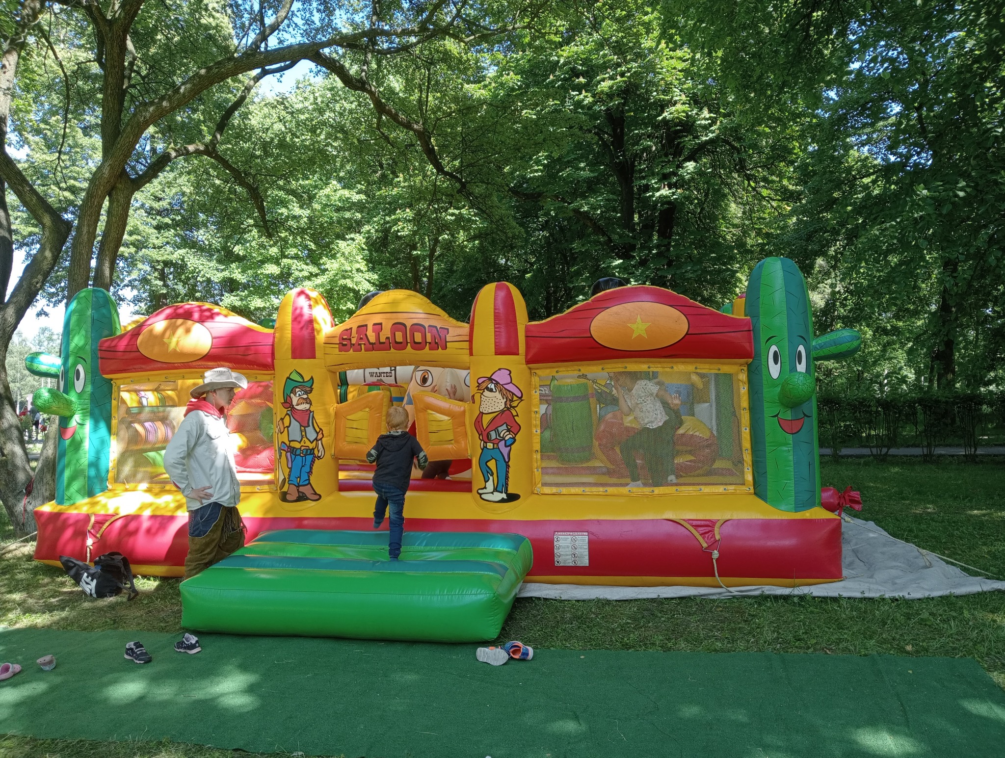 Kolorowy dmuchany zamek w stylu western 'Saloon' na trawie, z dzieckiem wspinającym się na wejście i mężczyzną w kapeluszu obserwującym. Tło stanowią drzewa.