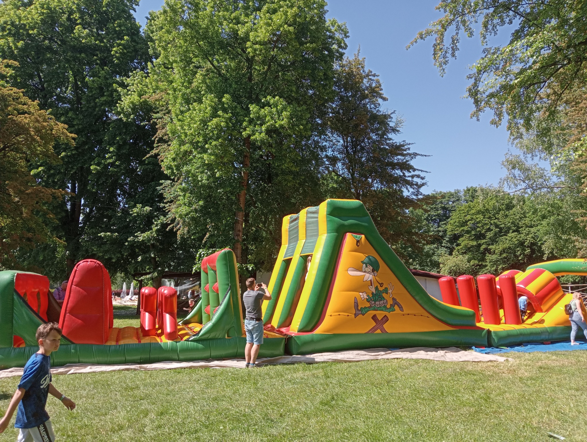 Kolorowy, rozłożysty dmuchaniec z motywem żołnierza na zielonym trawniku, otoczony drzewami. Osoba fotografuje atrakcję telefonem. Dziecko przechodzi obok.