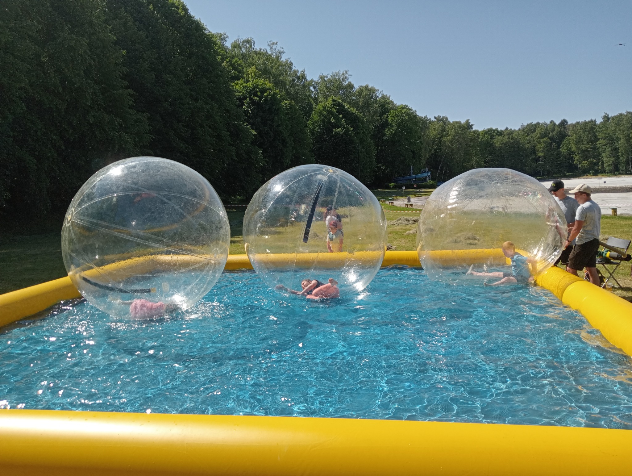 Trzy przezroczyste kule na wodzie w żółtym basenie, wewnątrz dzieci. W tle drzewa i obsługa. Atrakcja wodna na świeżym powietrzu, w słoneczny dzień.