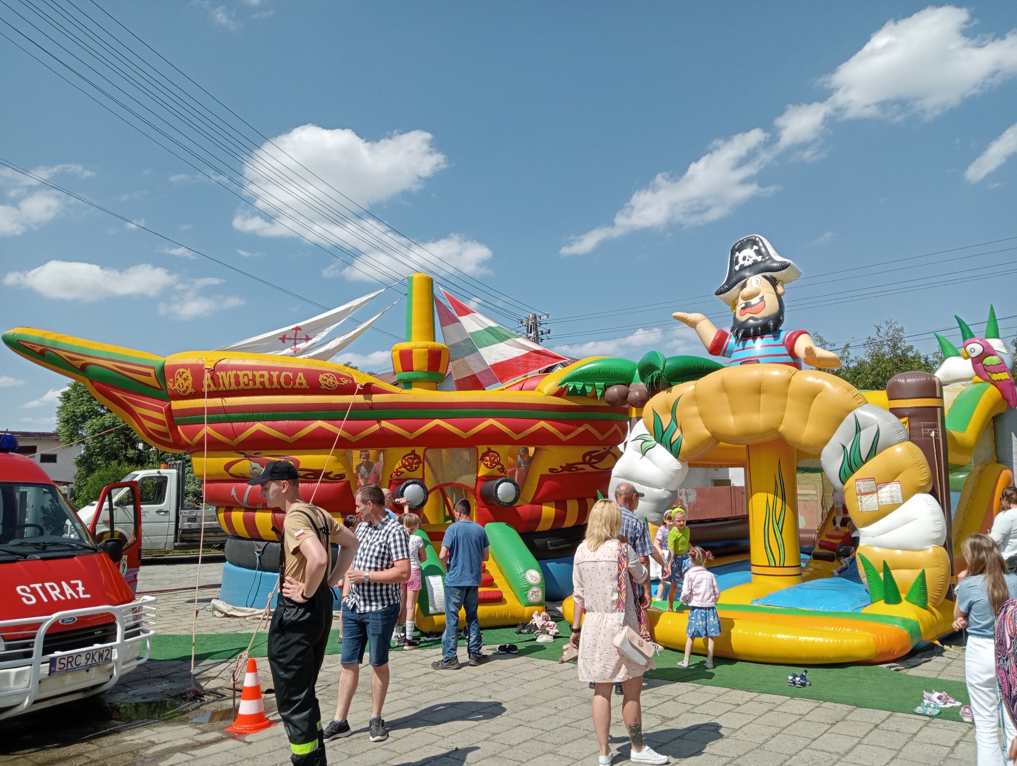 Kolorowy dmuchany zamek w kształcie statku pirackiego z animatorem w Rydułtowach. Dzieci bawią się w otoczeniu dorosłych. W tle wóz strażacki.