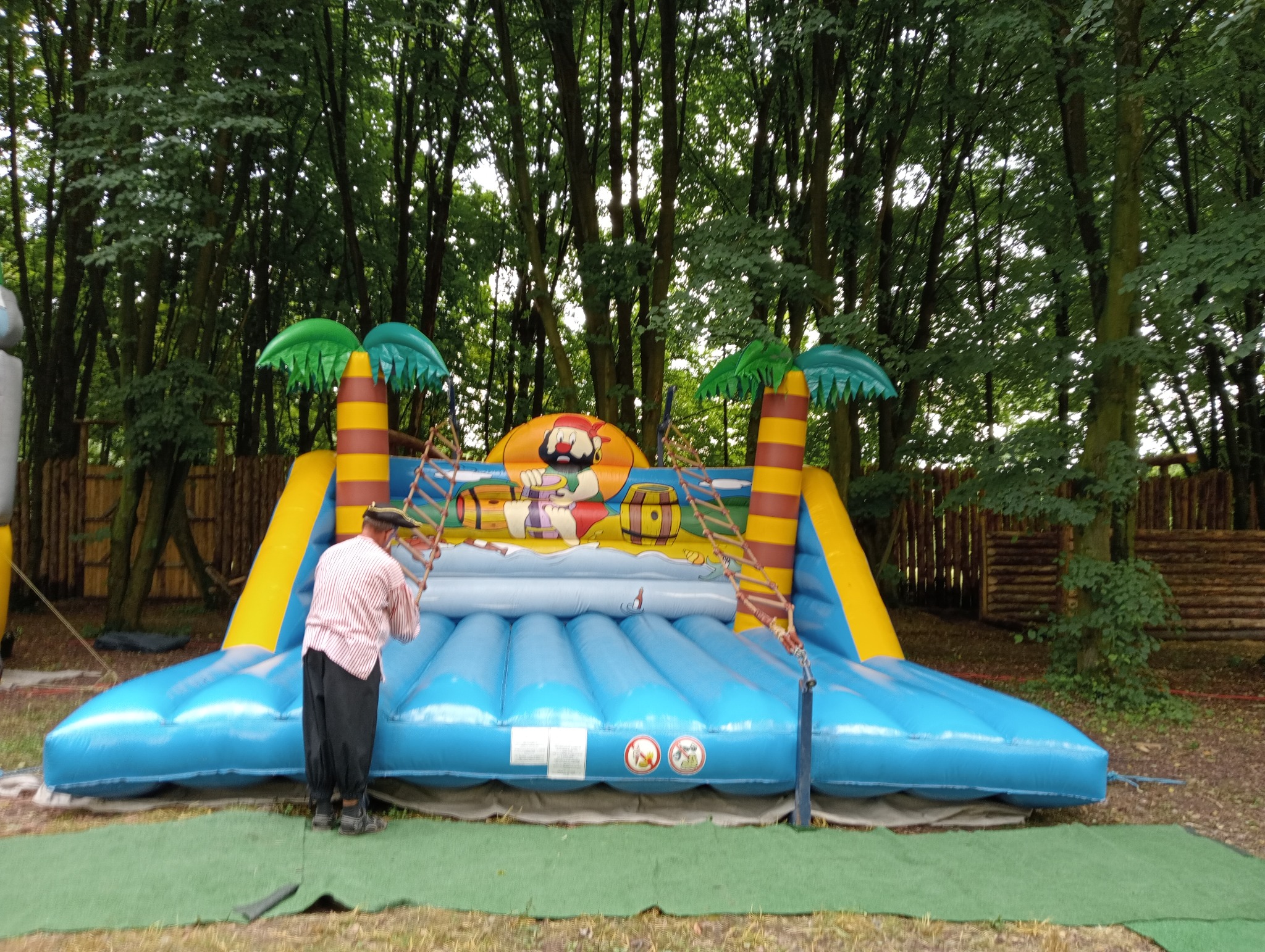 Kolorowy dmuchany zamek w pirackim stylu z palmami i zjeżdżalnią, ustawiony na trawie, przygotowywany przez mężczyznę w paski na tle drzew.