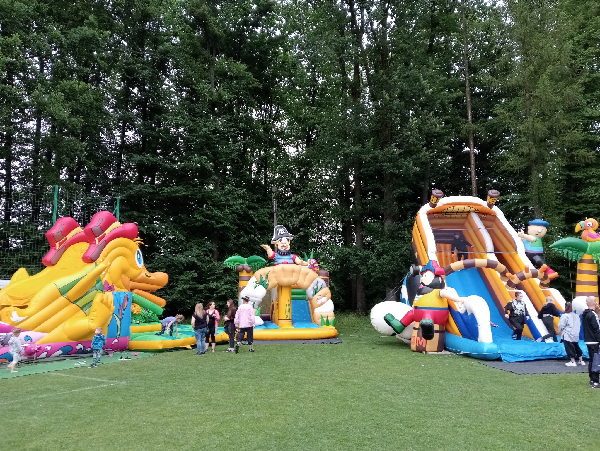 Kolorowe dmuchańce tematyczne na trawie, dzieci i dorośli korzystający z atrakcji. Tło stanowią drzewa, tworząc naturalne otoczenie imprezy plenerowej.