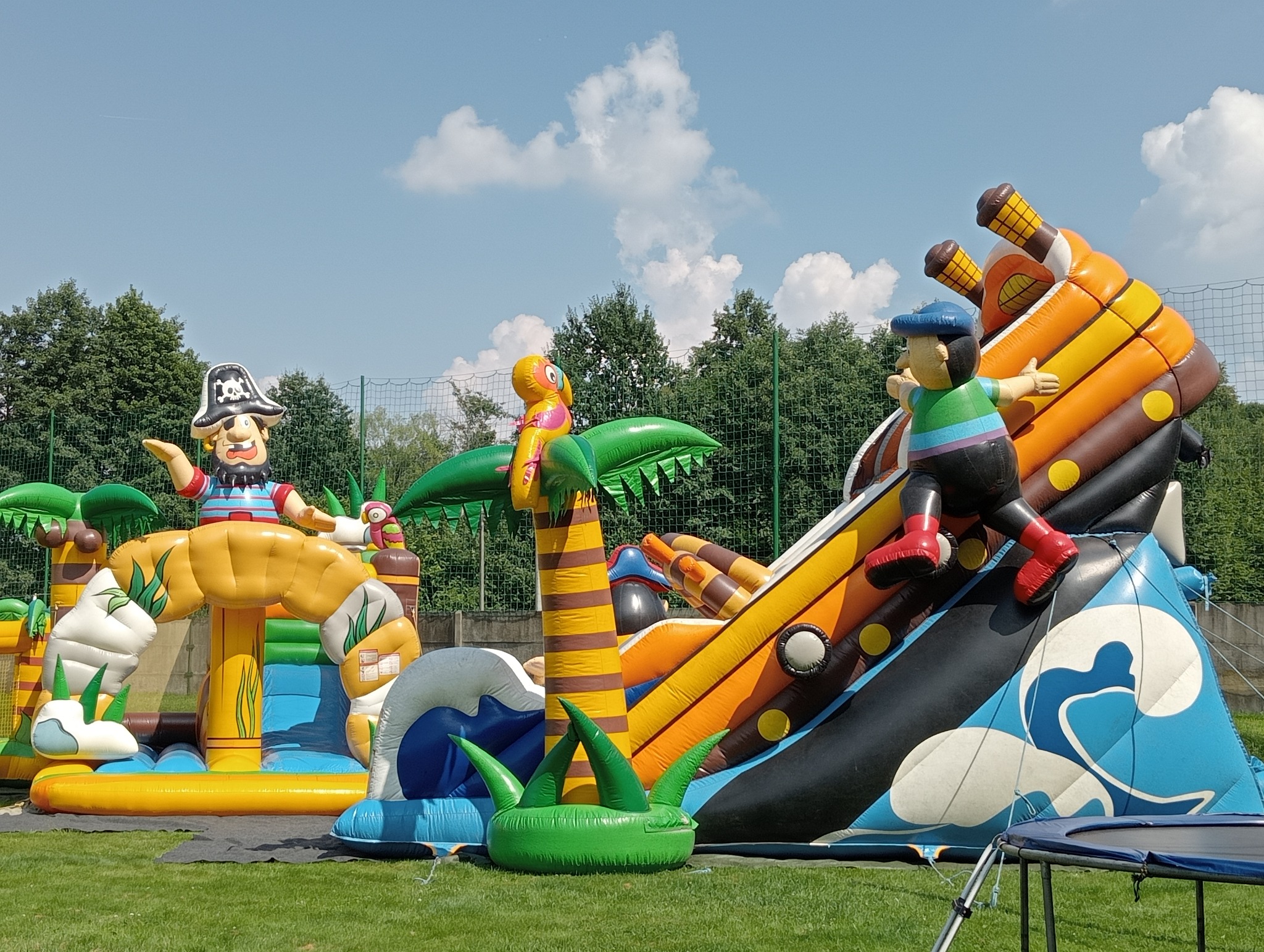 Kolorowy, rozbudowany dmuchany plac zabaw w pirackim stylu z postaciami piratów i palmami na trawiastym terenie, idealny na imprezy plenerowe dla dzieci.