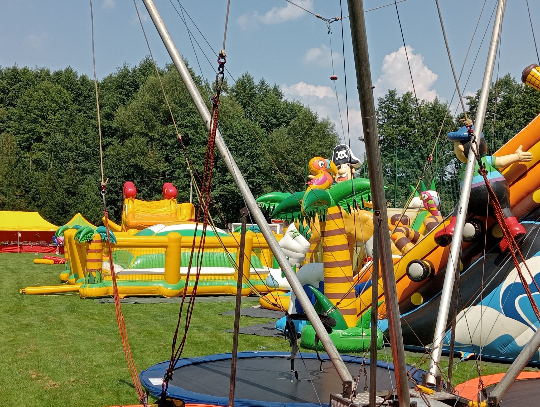 Kolorowy park dmuchańców na trawie, z trampoliną na pierwszym planie. W tle drzewa i błękitne niebo. Dmuchańce z motywami pirackimi i tropikalnymi.