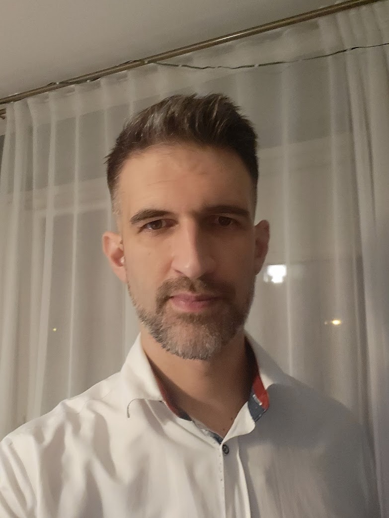 Mężczyzna w białej koszuli, portret od przodu, z krótką brodą i ciemnymi włosami z siwizną na tle białych zasłon.