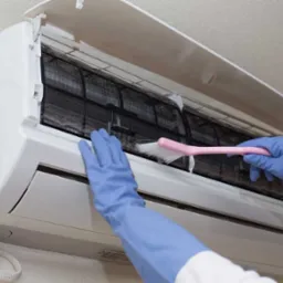 Czyszczenie filtra klimatyzacji ściennej z użyciem r&oacute;żowej szczotki. Dłonie w niebieskich rękawicach. Widoczny kabel zasilający. Otwarta obudowa klimatyzatora.