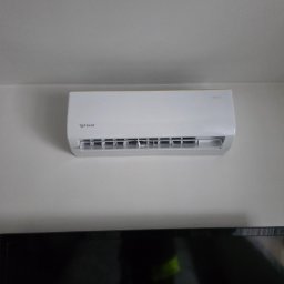 Montaż Klimatyzacji Mont-Instal Łukasz Szczepanek - Biały klimatyzator Rotenso zamontowany na ścianie nad czarnym telewizorem. Widoczne detale obudowy i wlotu powietrza. Minimalistyczne wnętrze, neutralne oświetlenie.