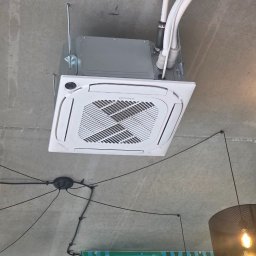 Montaż Klimatyzacji Mont-Instal Łukasz Szczepanek - Kasetonowa klimatyzacja Rotenso zamontowana na suficie, widoczne rury instalacyjne i fragment lampy. Surowy betonowy sufit. Ujęcie z dołu.