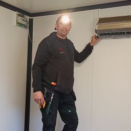 Montaż Klimatyzacji Mont-Instal Łukasz Szczepanek - Instalator z czołówką, w czarnym ubraniu roboczym, montuje klimatyzator w pomieszczeniu o białych ścianach. Widoczna skrzynka elektryczna i narzędzia.