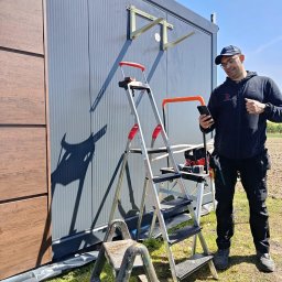 Montaż Klimatyzacji Mont-Instal Łukasz Szczepanek - Monolityczny budynek z zamontowanymi wspornikami klimatyzacji, instalator z telefonem i narzędziami na drabinie. Widok z zewnątrz, słoneczny dzień.