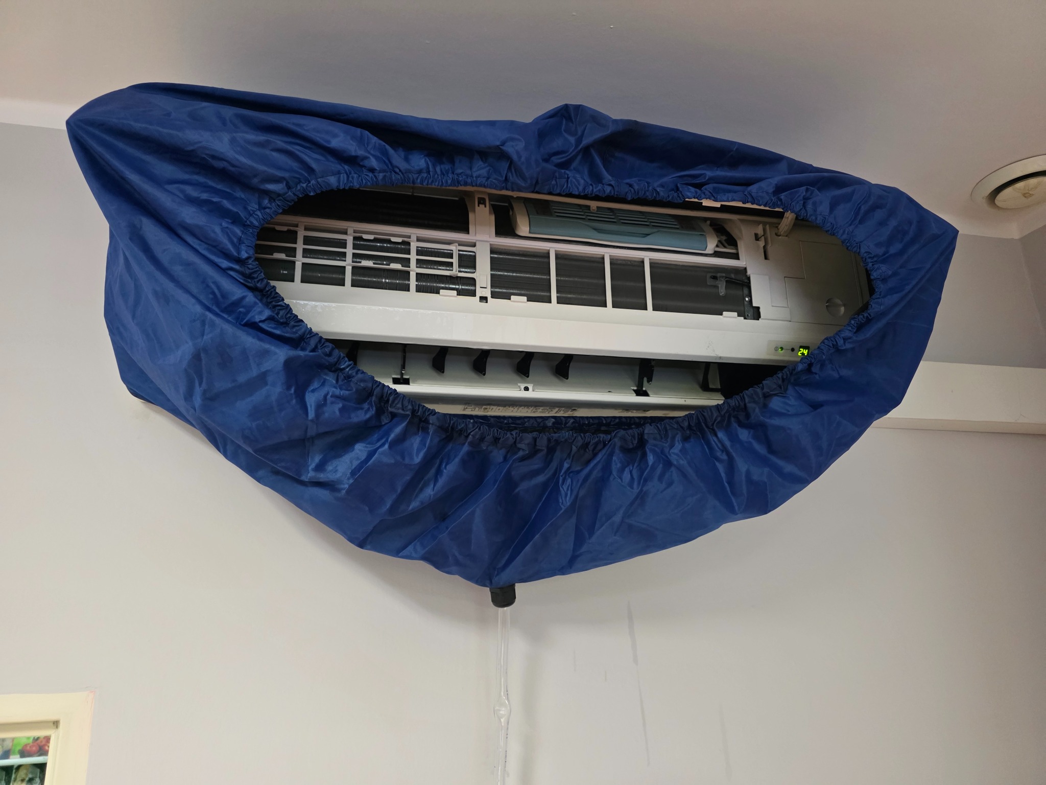 Klimatyzator ścienny w trakcie czyszczenia, zabezpieczony niebieskim pokrowcem z odprowadzeniem wody. Widoczne filtry i elementy wewnętrzne urządzenia.