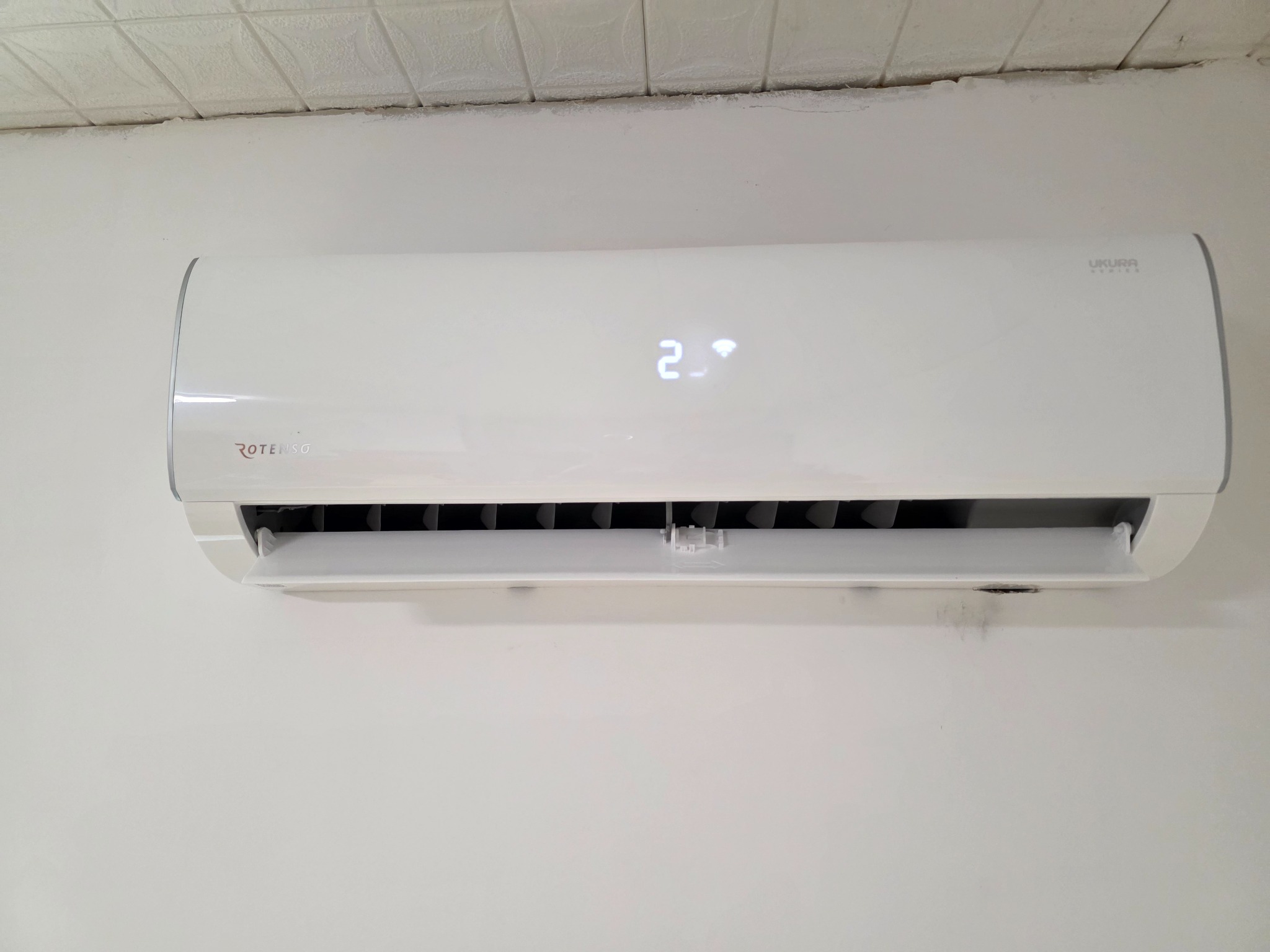 Biała klimatyzacja Rotenso Ukura Series zamontowana na ścianie, wyświetlająca temperaturę 21 stopni Celsjusza, widok z przodu, wnętrze pomieszczenia.