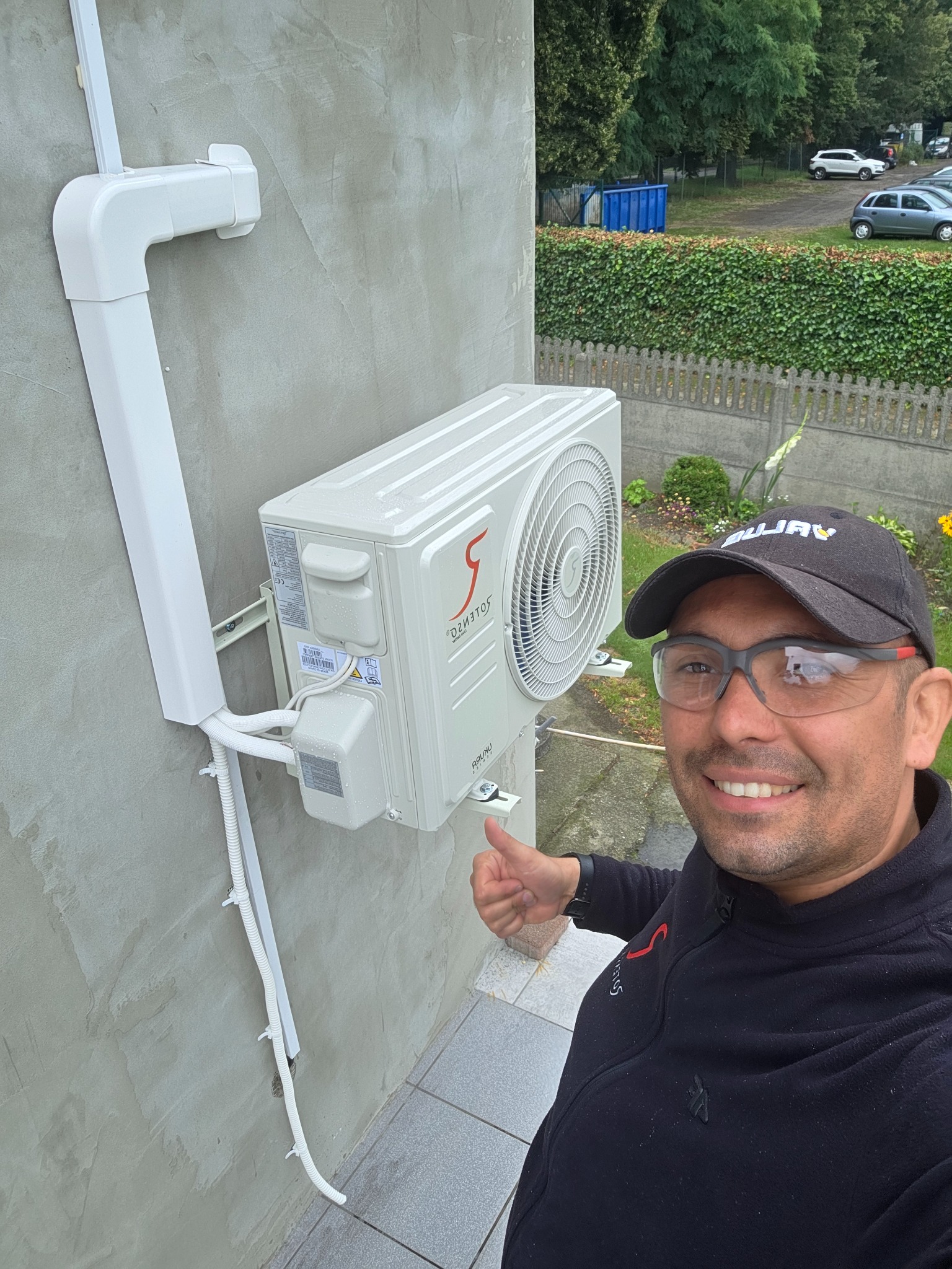 Biały klimatyzator zewnętrzny zamontowany na szarej ścianie, z białymi rurami osłonowymi. Instalator uśmiechnięty, z kciukiem w górę, w czapce i okularach ochronnych.