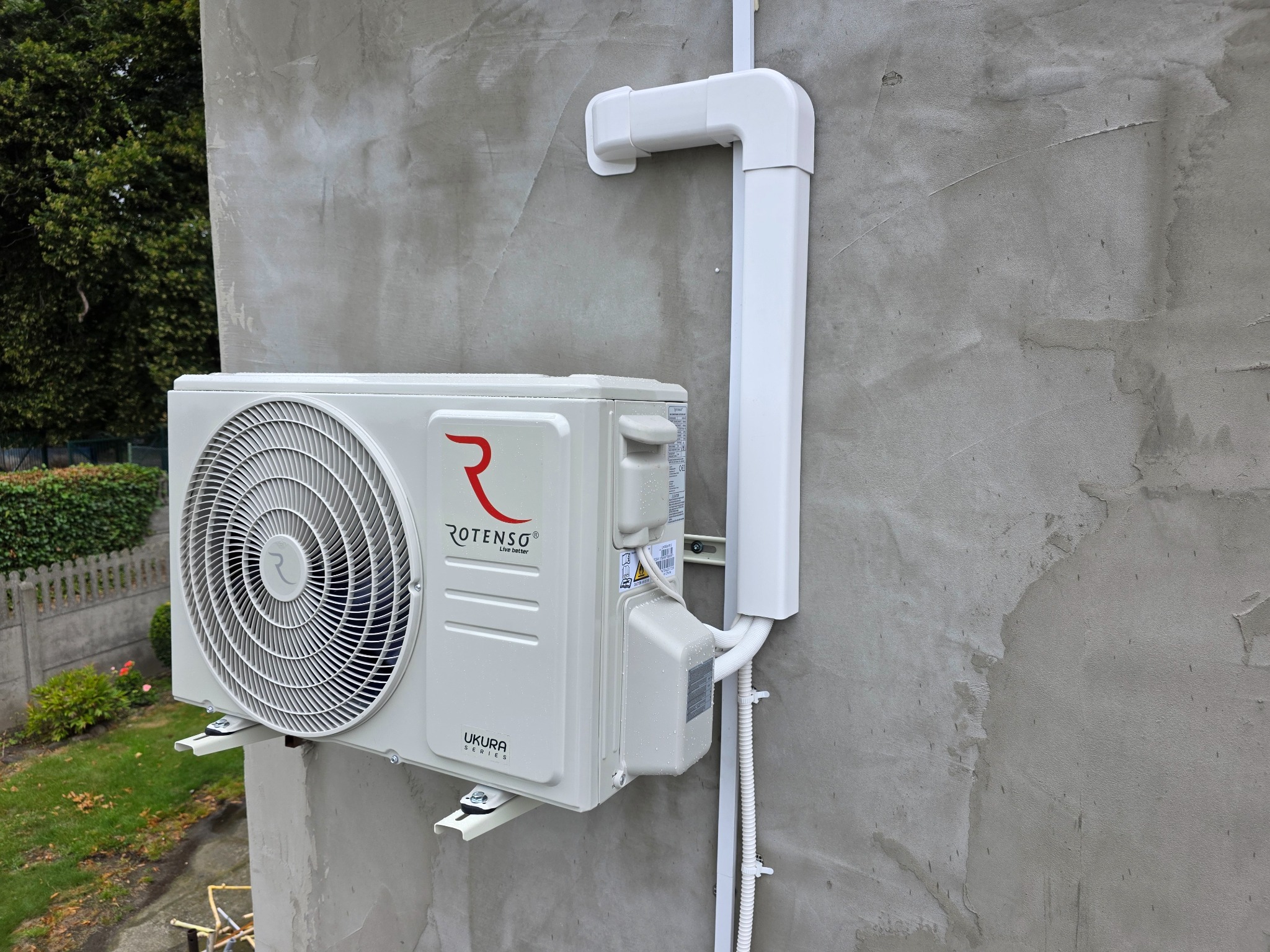 Zamontowana jednostka zewnętrzna klimatyzacji Rotenso Ukura na szarej ścianie budynku, z widocznymi rurami odprowadzającymi. Tło: zieleń ogrodu.