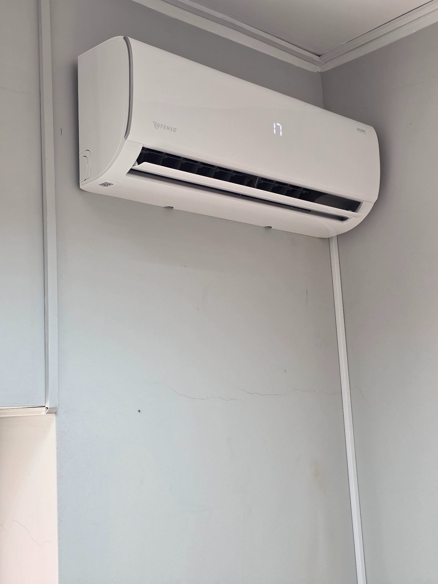 Biała klimatyzacja ROTENSO UKURA zamontowana na szarej ścianie, wyświetlająca temperaturę 17 stopni, widok z dołu, oświetlenie naturalne.