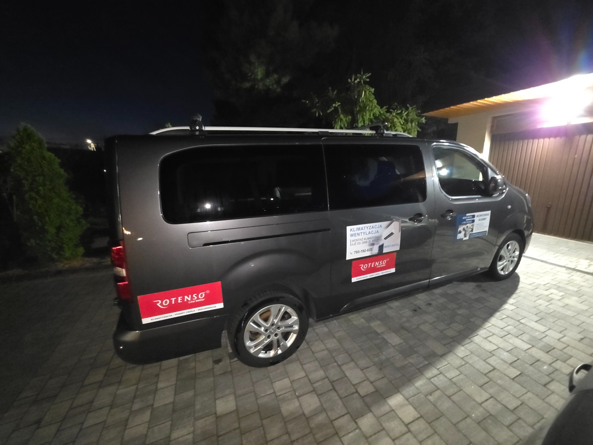 Szary van firmy Rotenso z reklamą klimatyzacji i wentylacji, zaparkowany nocą na brukowanym podjeździe przed garażem. Widoczne logo i dane kontaktowe firmy.
