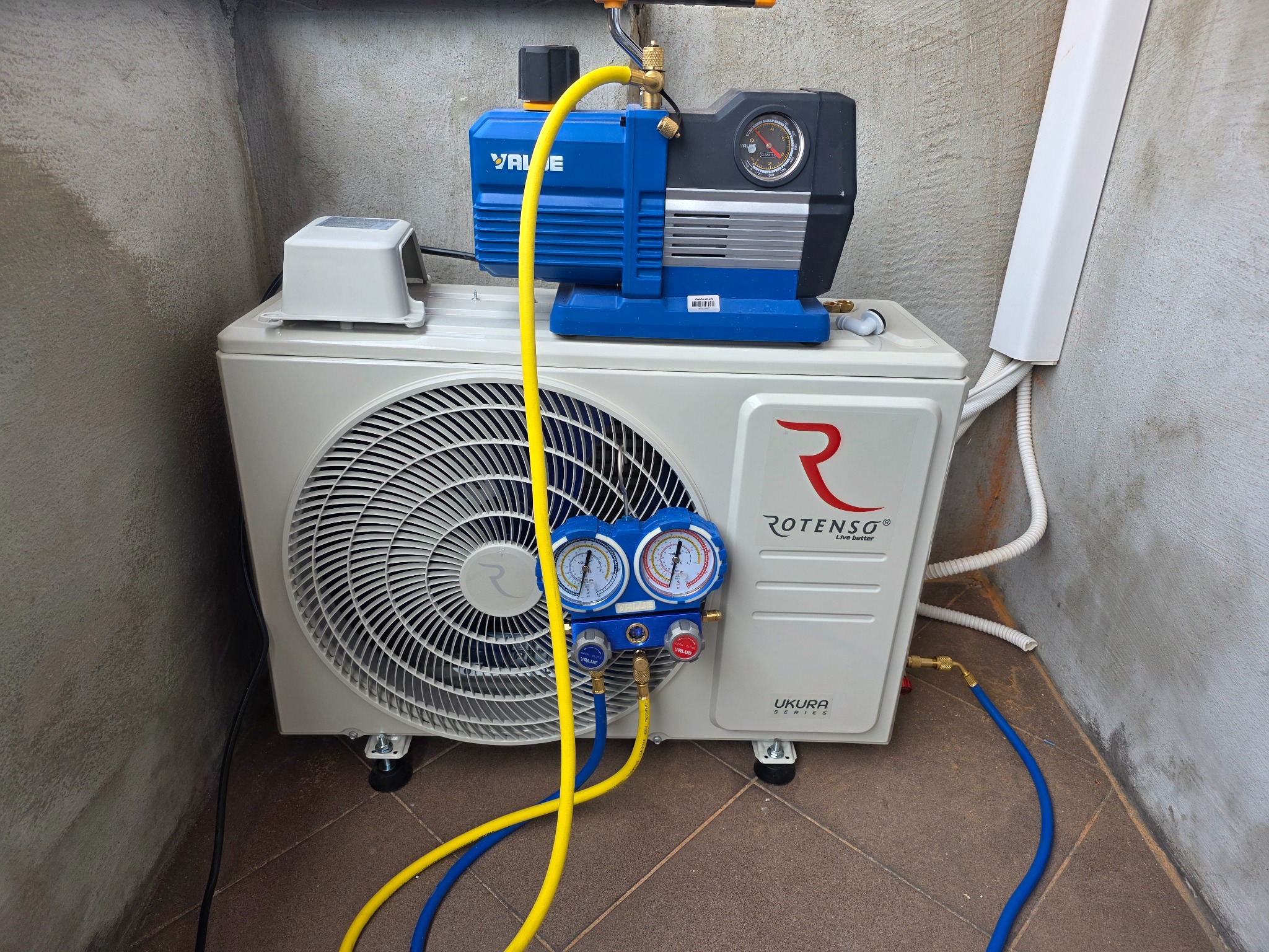Biała jednostka zewnętrzna klimatyzacji Rotenso z podłączonymi manometrami i pompą próżniową Value podczas serwisu w pomieszczeniu technicznym.
