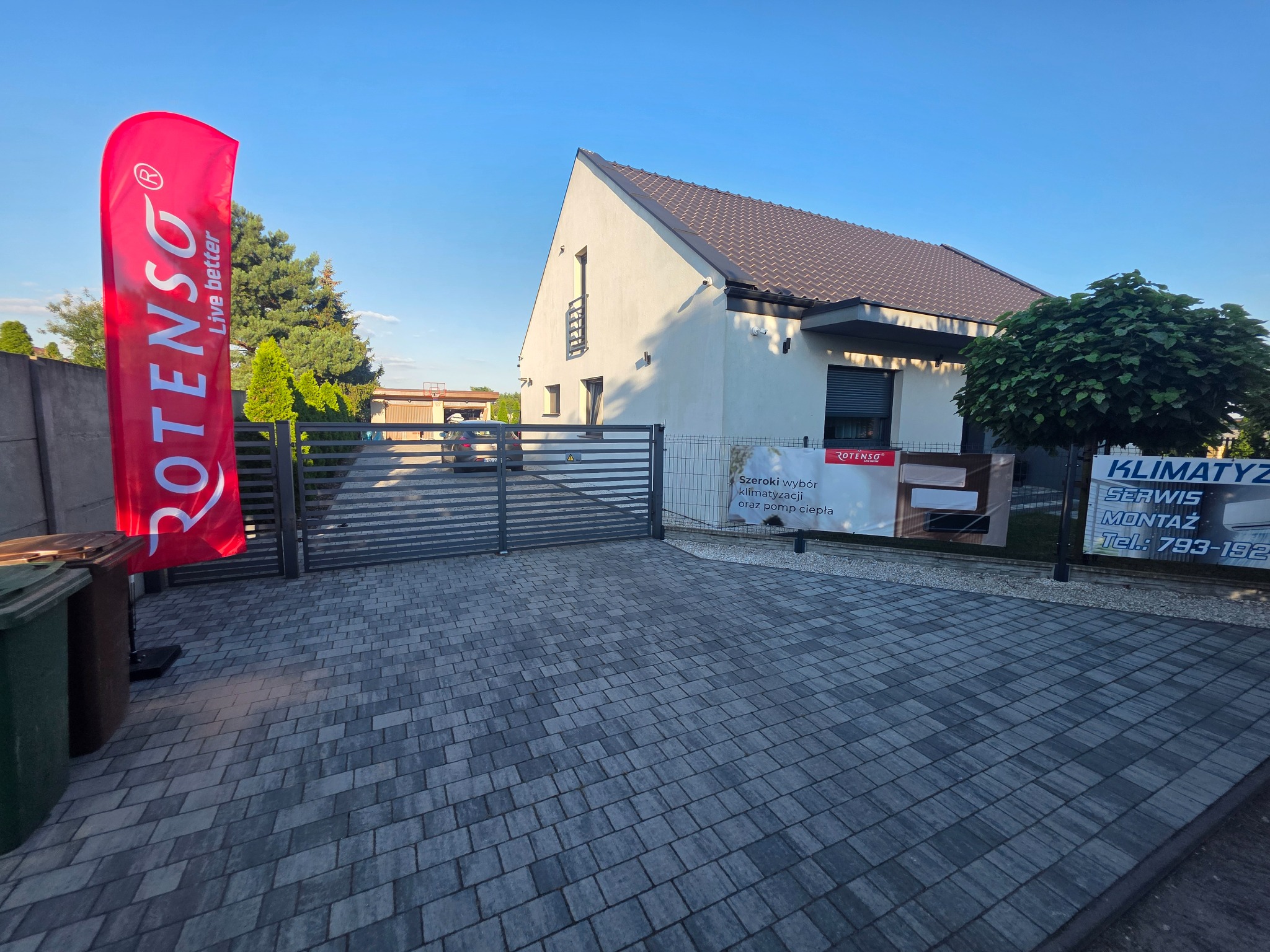 Nowoczesny dom jednorodzinny z szarą kostką brukową na podjeździe, brama wjazdowa, reklama firmy Rotenso oferującej klimatyzację i pompy ciepła, błękitne niebo.