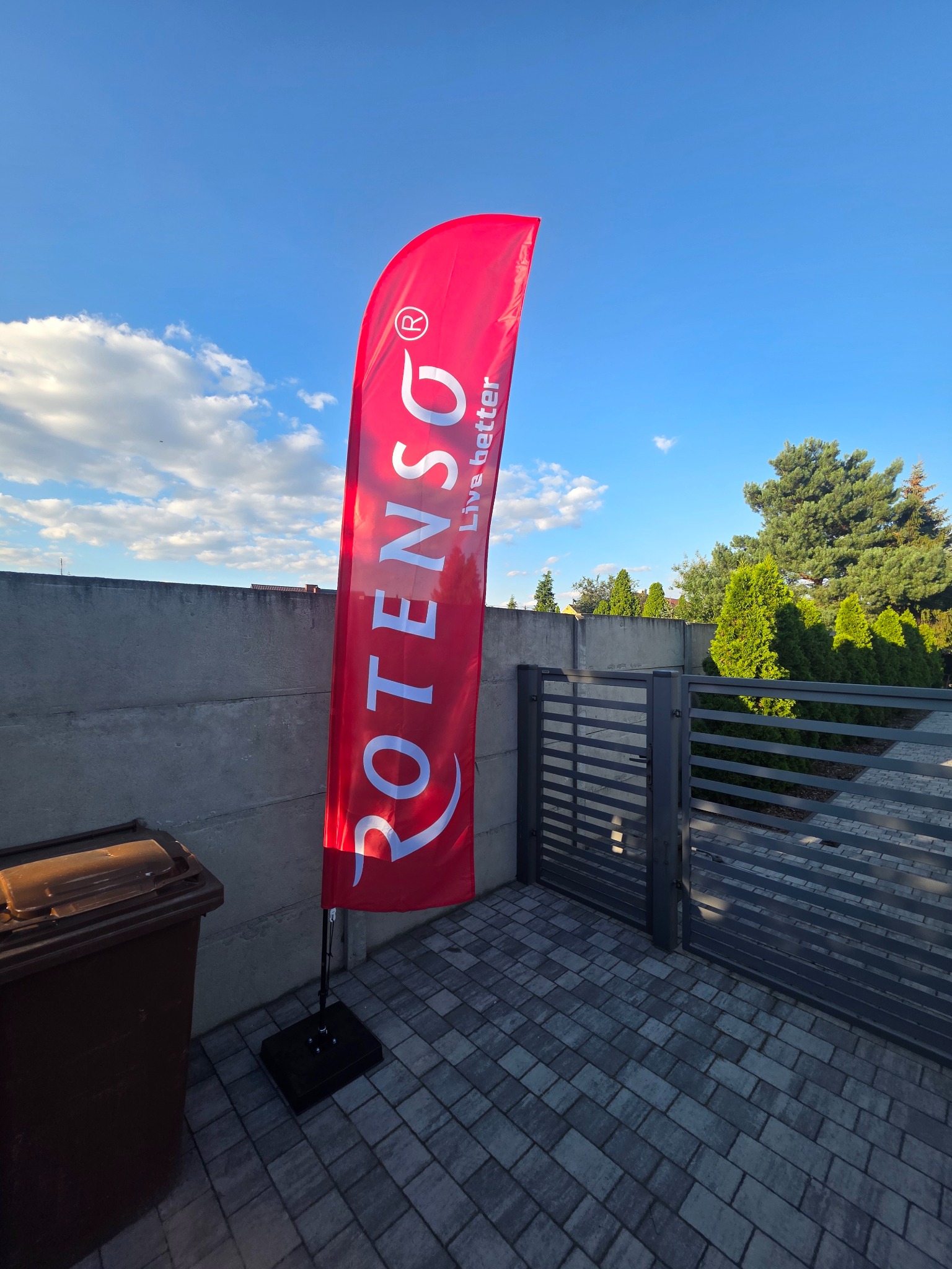 Czerwony, pionowy baner reklamowy firmy Rotenso z napisem „Live better” na tle błękitnego nieba, szarego muru i zielonych drzew. Baner stoi na kostce brukowej obok brązowego kosza.