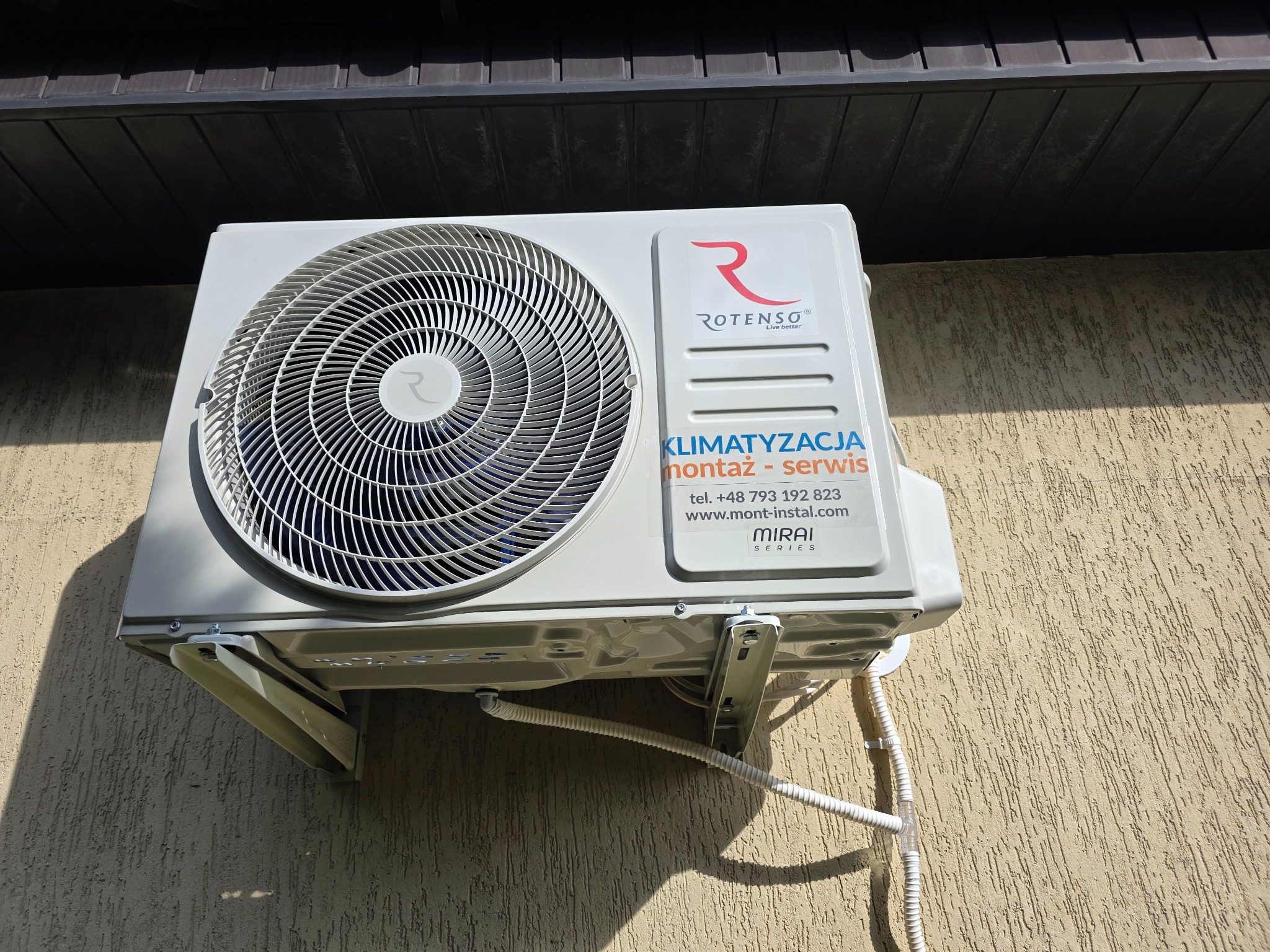 Biała jednostka zewnętrzna klimatyzacji Rotenso zamontowana na elewacji budynku. Widoczne przewody i logo firmy. Kąt ujęcia z dołu podkreśla solidność montażu.