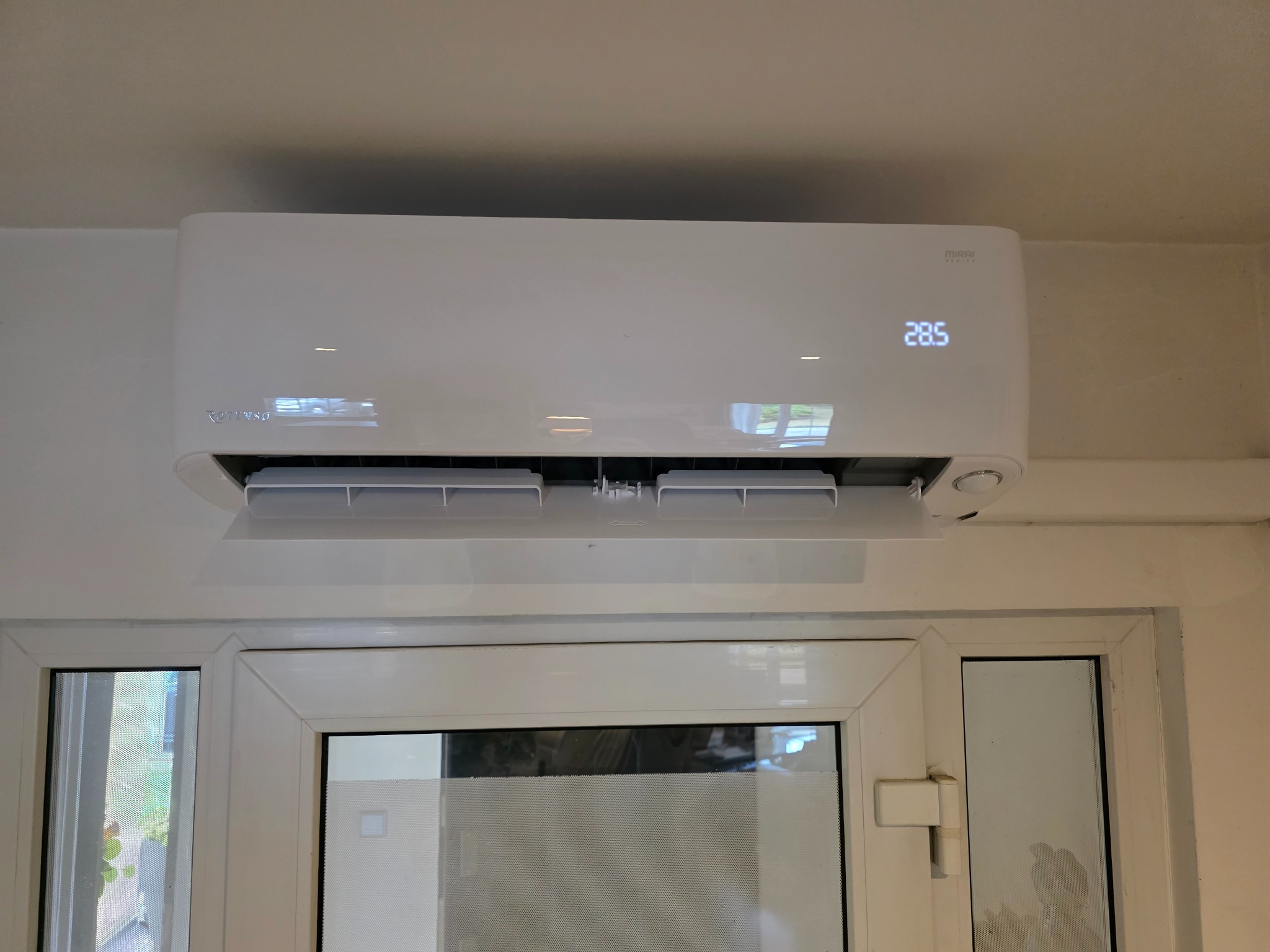 Biała klimatyzacja typu split, model Mirai, zamontowana nad oknem. Widoczny wyświetlacz z temperaturą 28.5 stopni. Minimalistyczny design i jasne otoczenie.