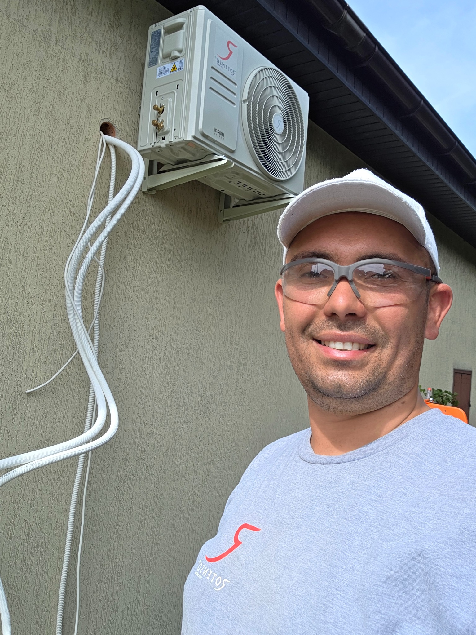 Zamontowana klimatyzacja na elewacji budynku, widoczne wystające kable, na pierwszym planie uśmiechnięty monter w okularach ochronnych i firmowej koszulce.