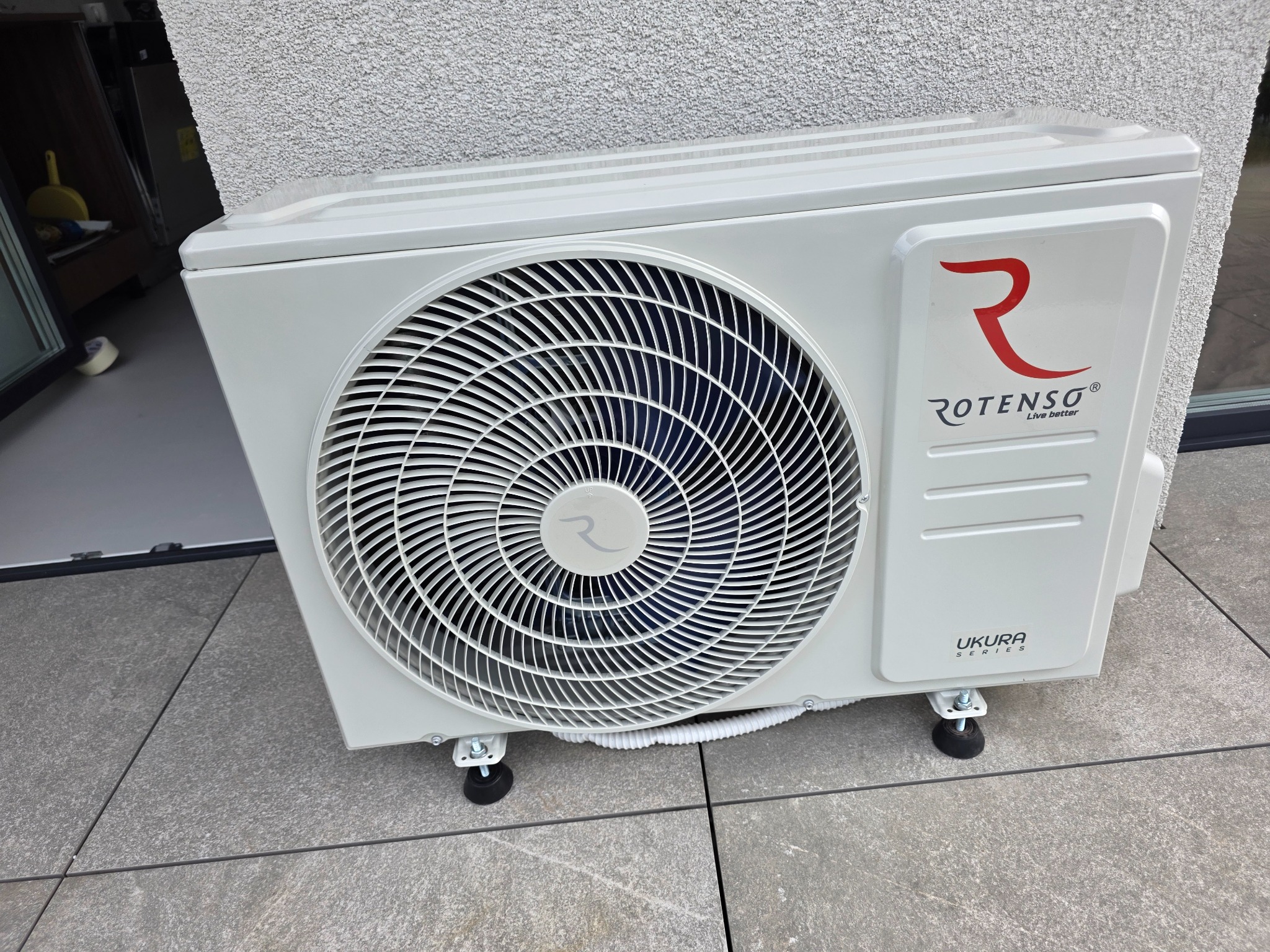 Biała jednostka zewnętrzna klimatyzacji Rotenso Ukura Series, zamontowana na szarym tarasie przy ścianie budynku. Widoczne logo i detale montażowe.
