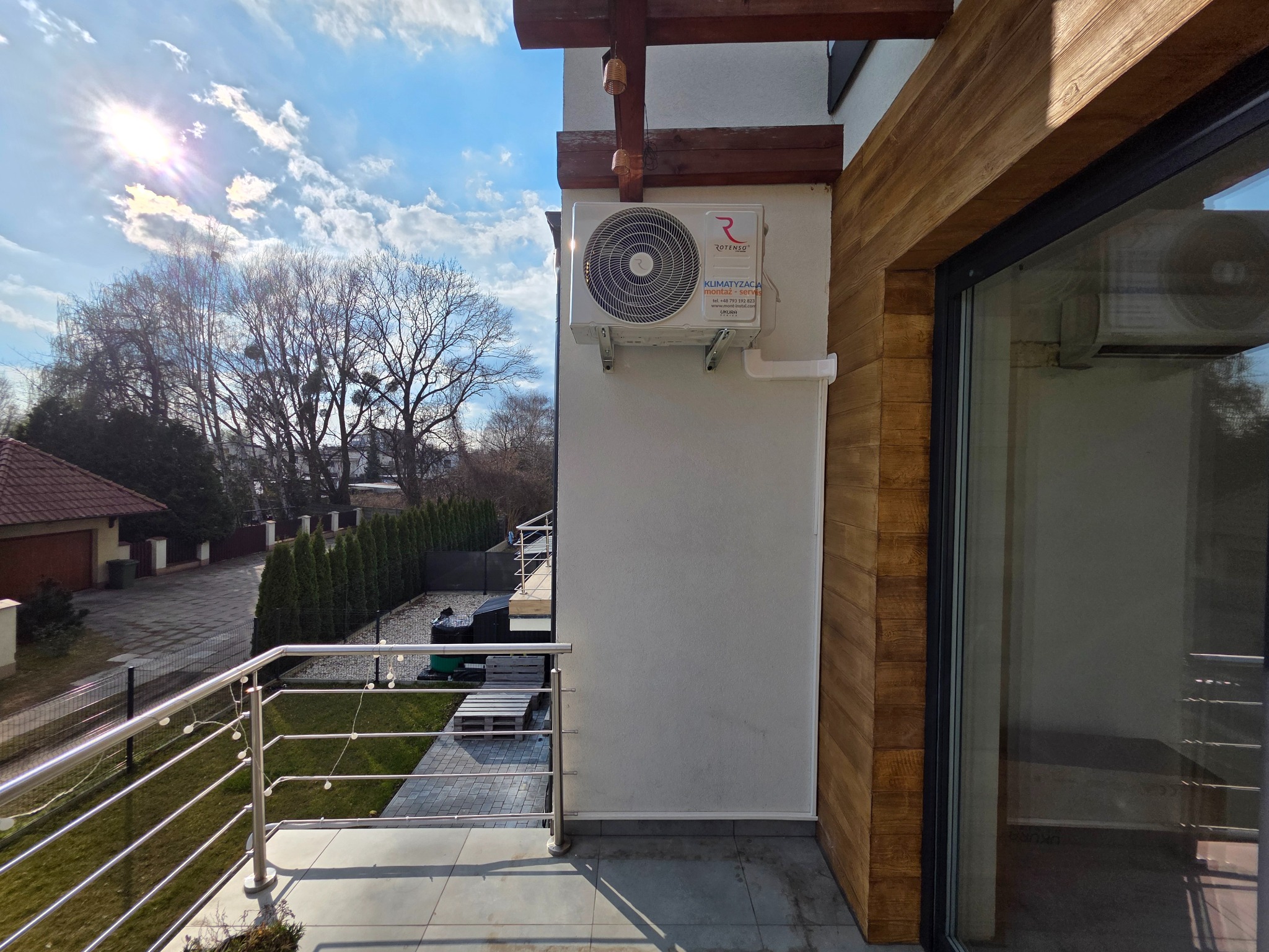 Biała jednostka klimatyzacji Rotenso zamontowana na ścianie budynku, widoczna z balkonu z metalową balustradą w słoneczny dzień. Widok na okolicę.