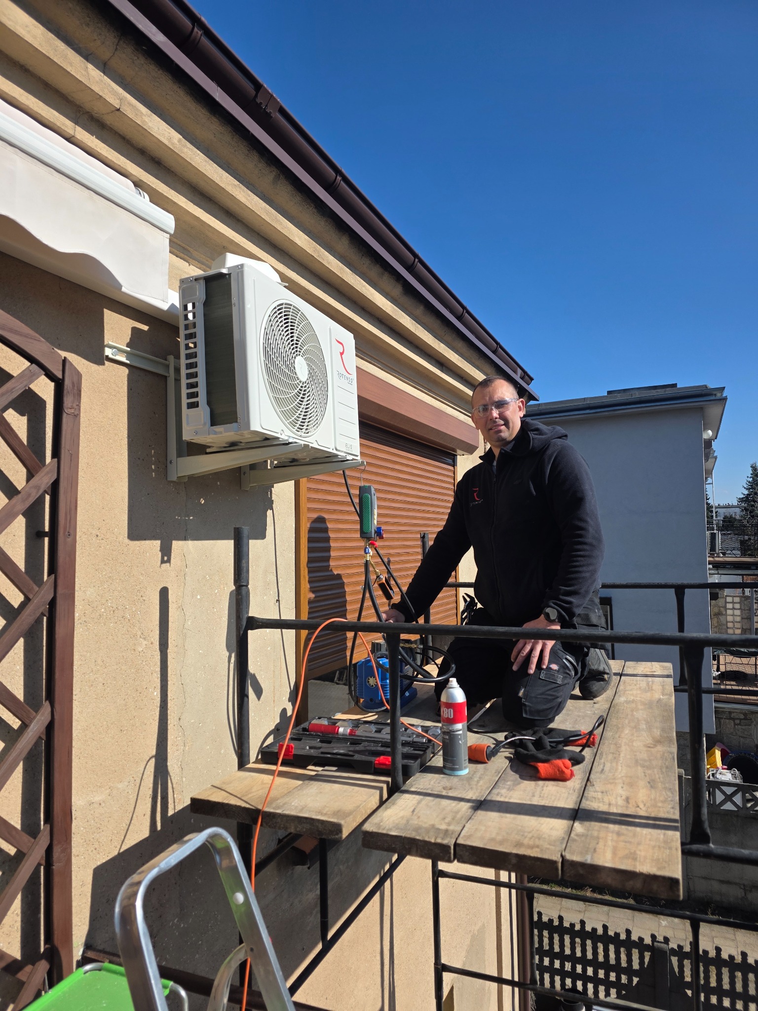 Montaż klimatyzatora na elewacji budynku. Instalator na podeście roboczym, narzędzia obok. Widoczne elementy instalacji i jednostka zewnętrzna klimatyzacji.